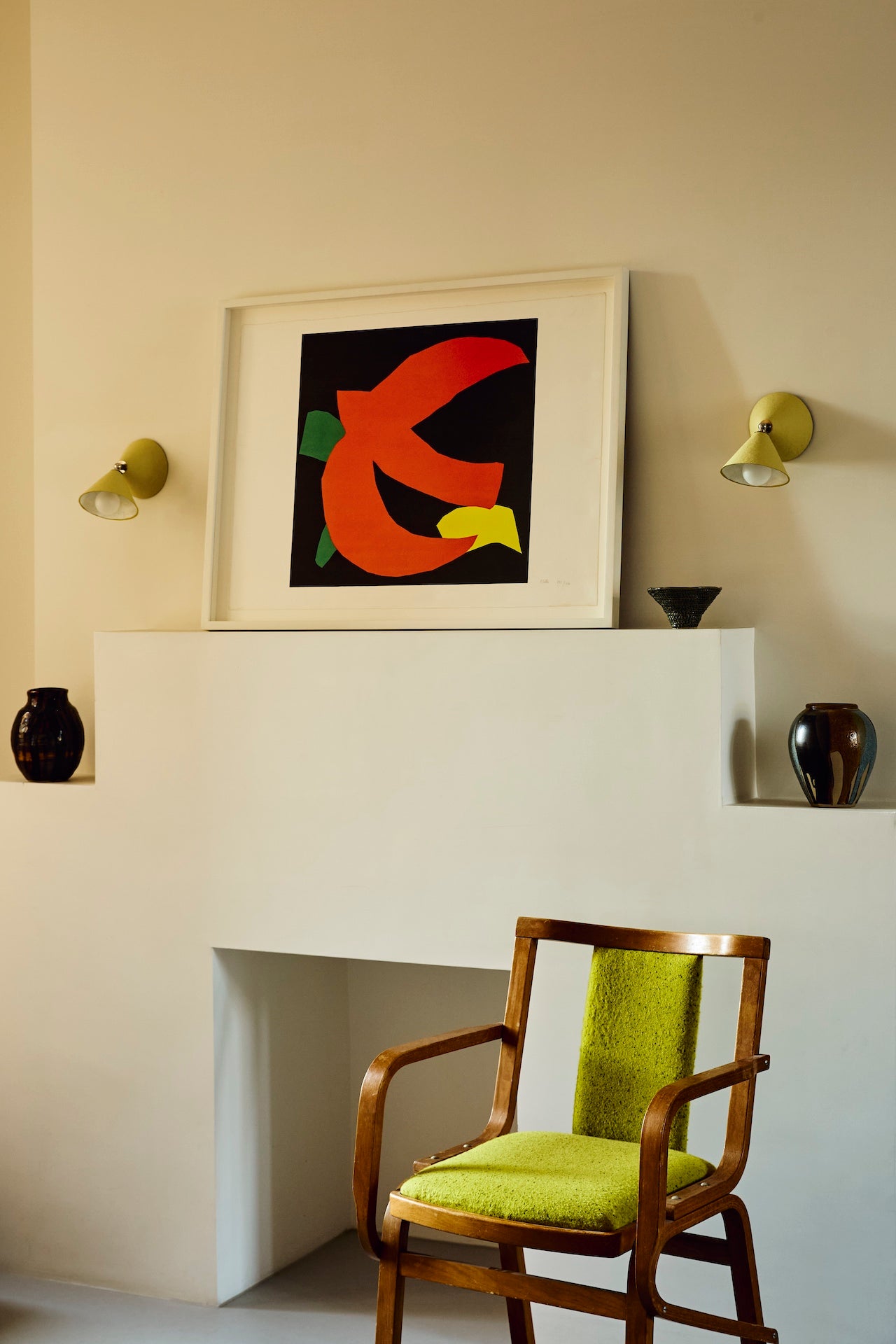 Lifestyle image of two hardwired Rotor wall lights- conical shape, hand-painted in light yellow colour with nickel finishes shown attached to the wall on either side of a Philip Sutton picture, above a mantel, holding decorative ceramics, with a chair in the foreground.