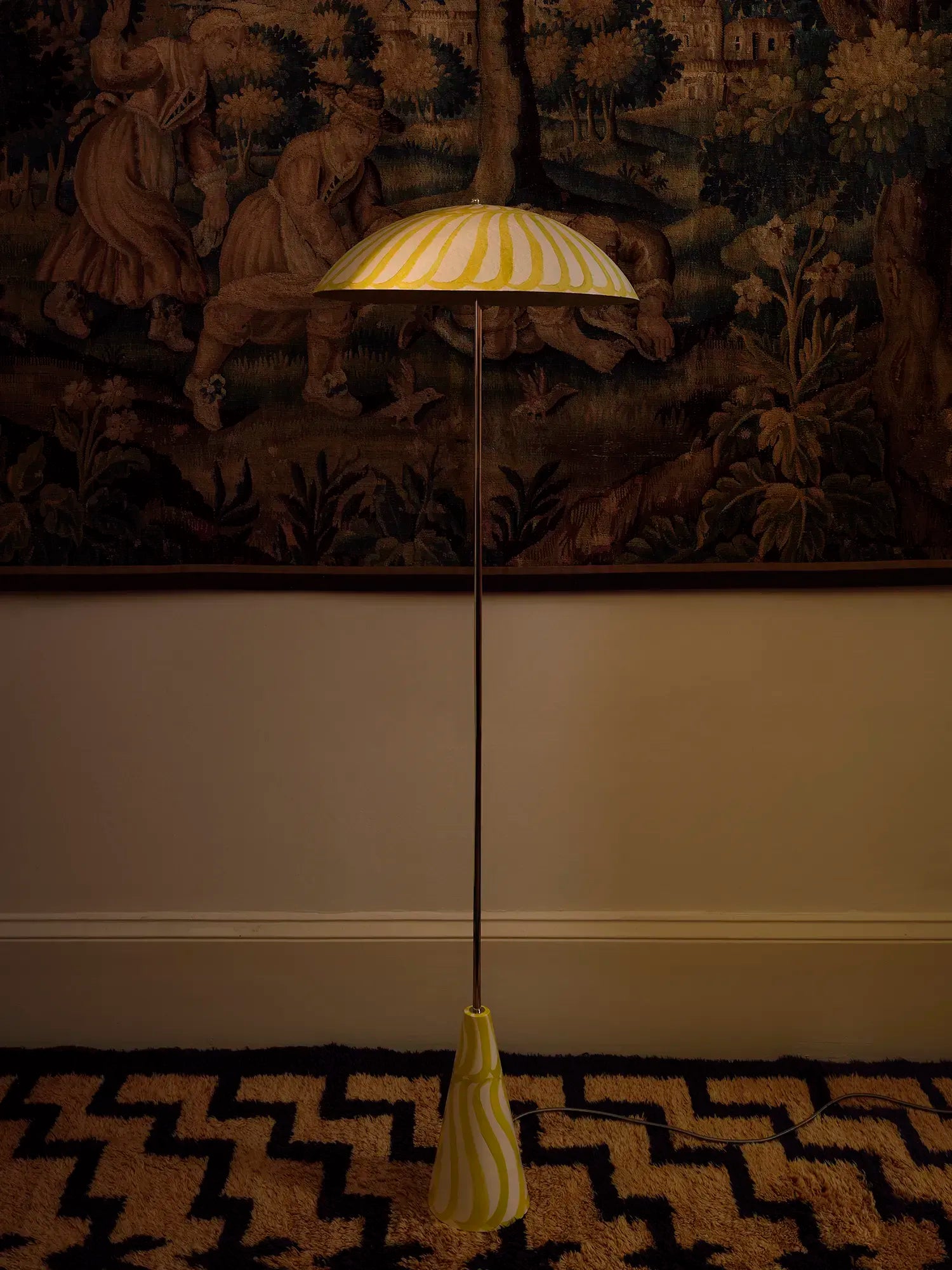 A vintage-style floor lamp with a striped yellow and white dome shade and a matching base, standing on a patterned rug in front of a dark tapestry.