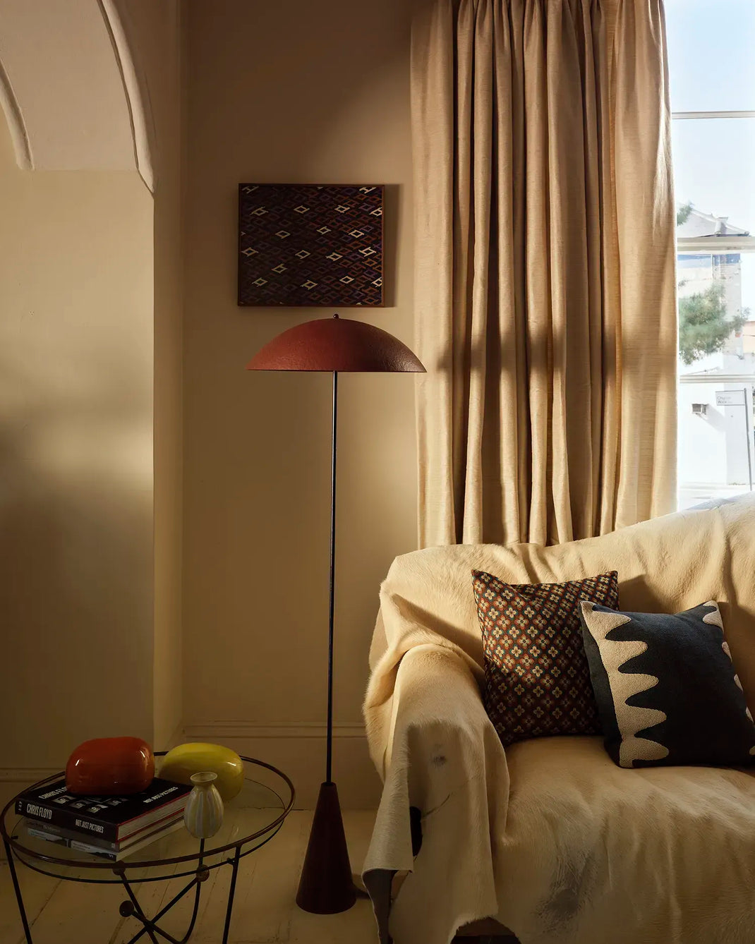 A cozy interior scene with a cream sofa draped in a throw, patterned pillows, a floor lamp with a dome shade, beige curtains, and a small side table with books and decorative objects.