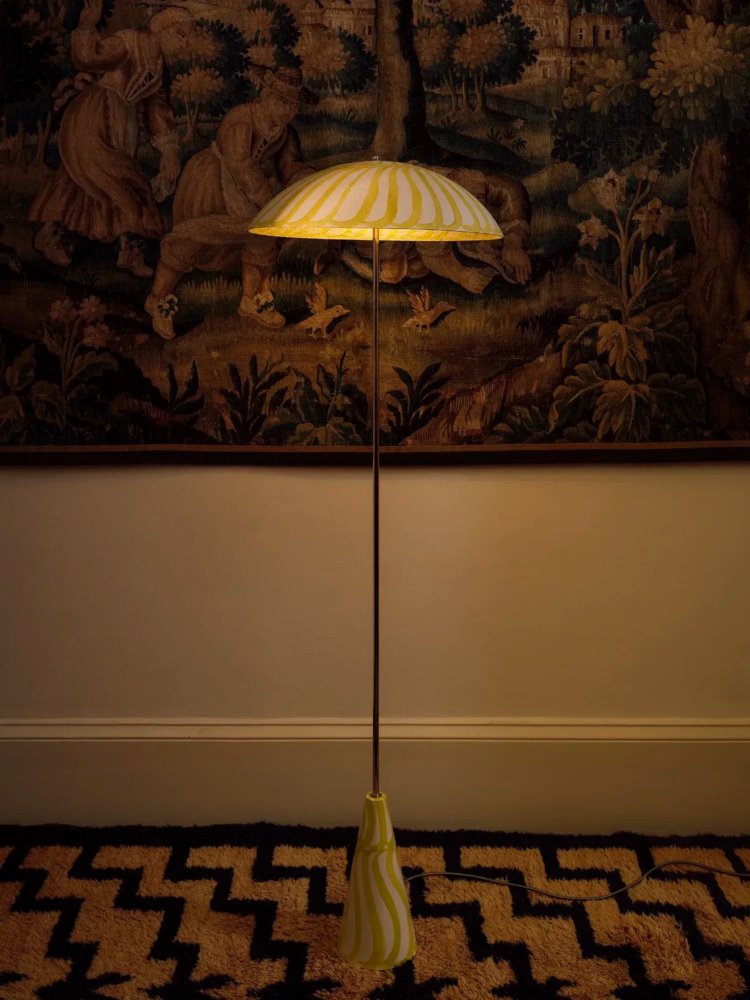 A vintage-style floor lamp with a striped cream and yellow shade, a slim dark metal pole, and a matching base, standing on a patterned rug in front of a tapestry.