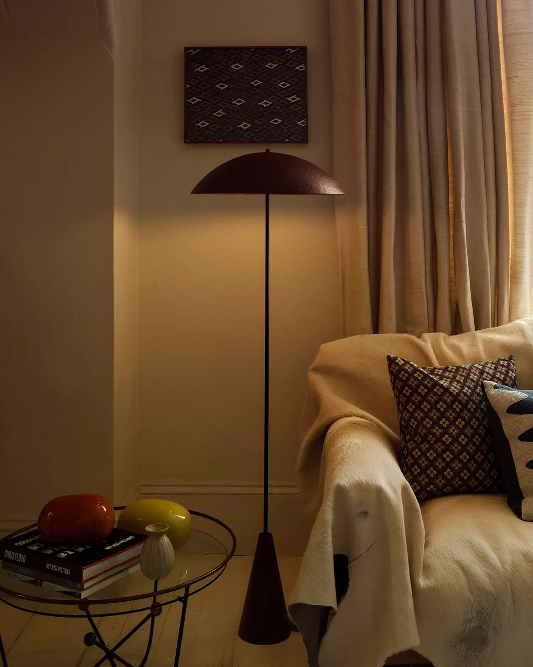 A mid-century modern floor lamp with an oxblood dome shade and tapered base stands in a warmly lit living room next to a cream sofa with patterned pillows.