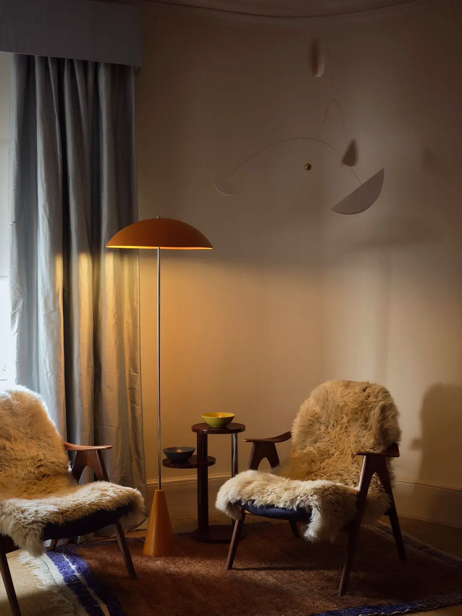 A warm, moody interior scene with two wooden chairs draped in white sheepskin, a floor lamp with a dome shade, a small side table, and a textured wall in a dimly lit room.