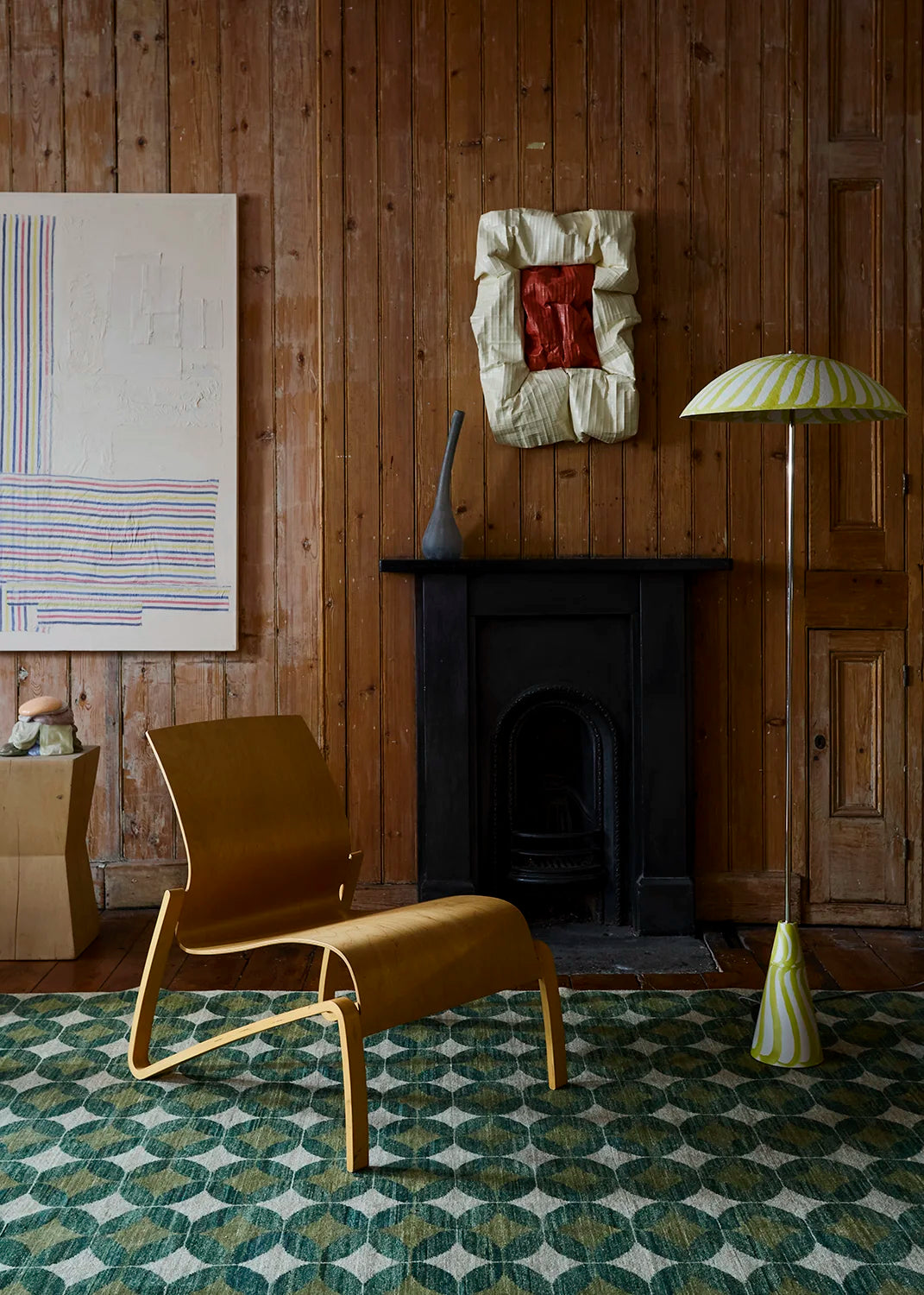 A mid-century modern wooden lounge chair in a warm tan finish, positioned in a rustic room with wood-paneled walls, a black fireplace, a green and white patterned rug, and a striped floor lamp.