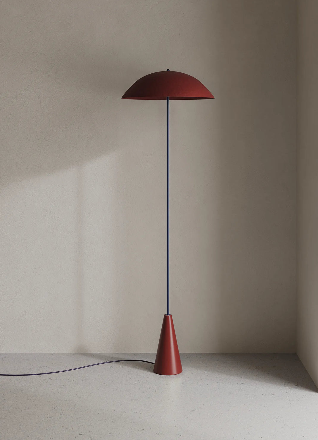 A modern floor lamp with a matte oxblood dome shade and base, supported by a thin dark blue pole, standing on a light gray floor against a beige wall.