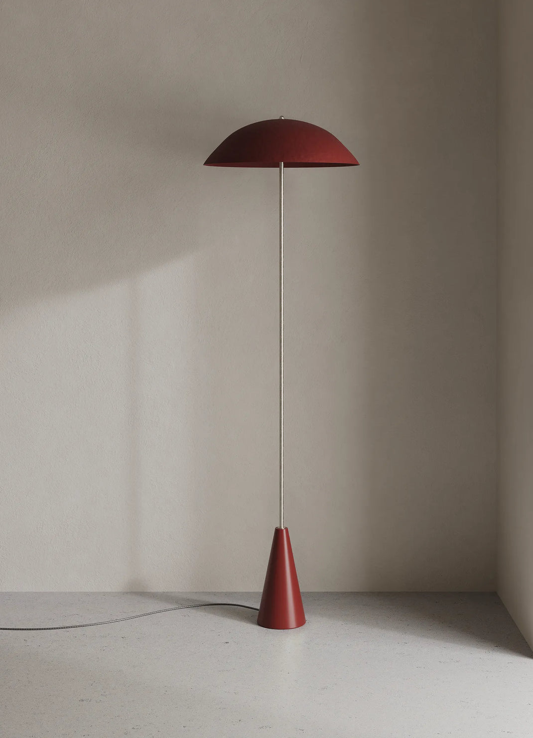 A modern floor lamp with an oxblood dome-shaped shade and a matching cone base, standing on a light gray floor against a textured beige wall.
