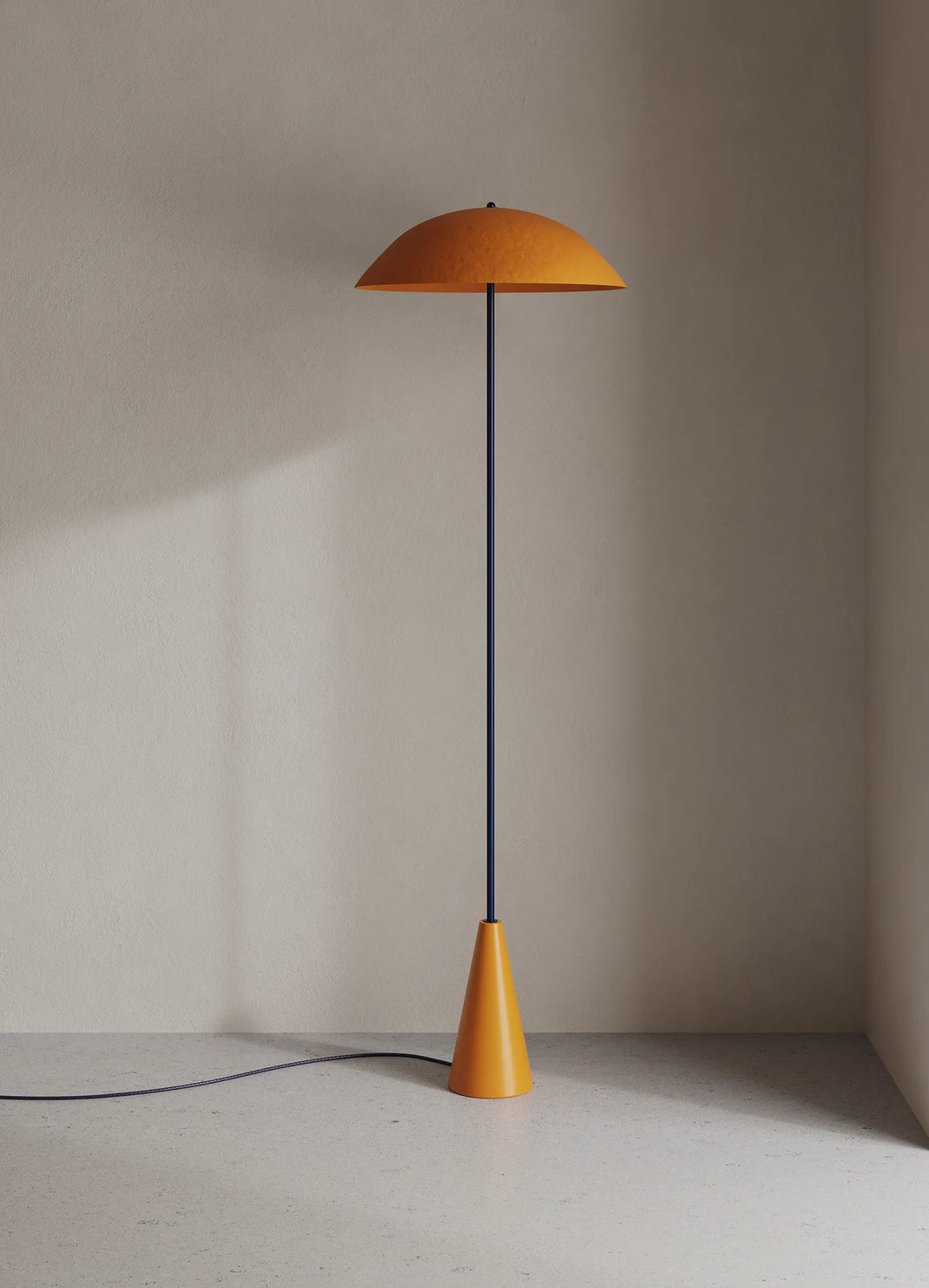A modern floor lamp with a wide, dome-shaped orange shade, a thin dark blue central pole, and a tapered orange base, standing on a light gray floor against a textured beige wall.