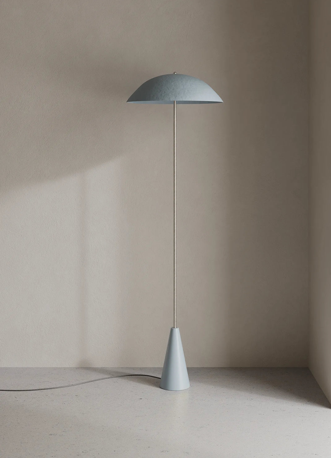 A minimalist floor lamp with a wide, shallow dome shade, a slim central stem, and a tapered base, shown in a pale blue-color against a warm beige wall and floor.