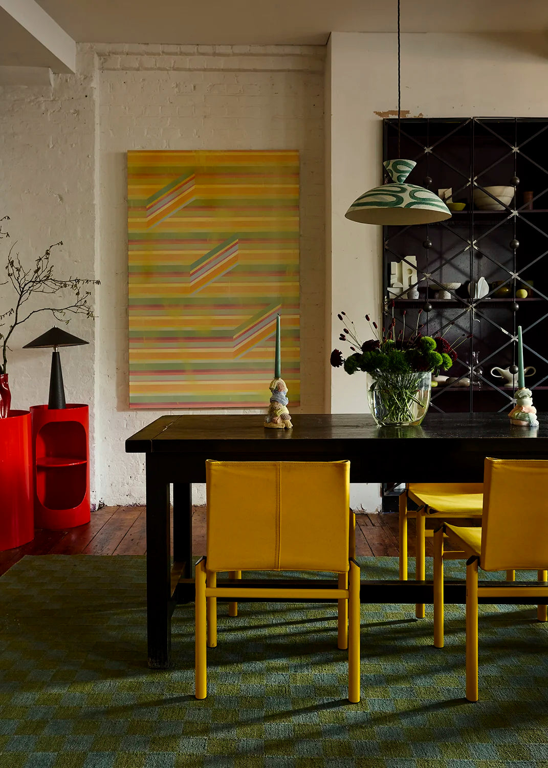 A dining room scene with a green and white pendant hanging above a dark rectangular table and four mustard yellow chairs, set on a green patterned rug. A large abstract striped painting hangs on the wall, and a black shelving unit stands to the right.