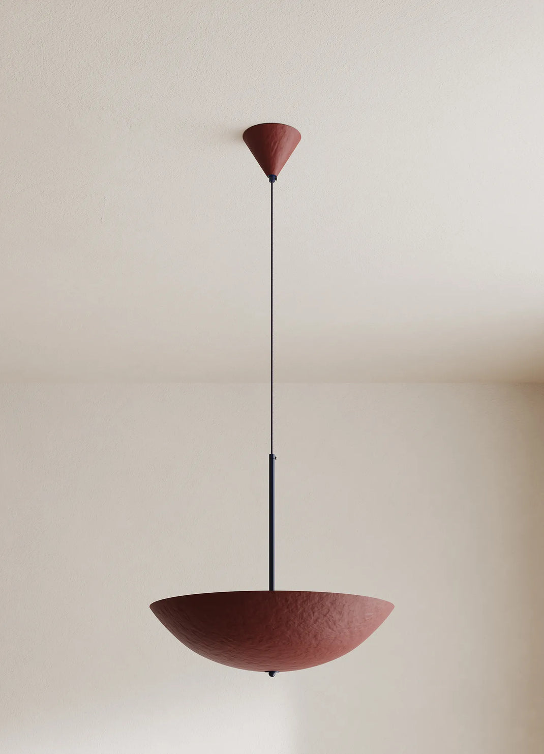A modern pendant light with a textured deep red-colored bowl shade and a thin dark blue suspension rod, shown hanging against a neutral cream-colored wall and ceiling.