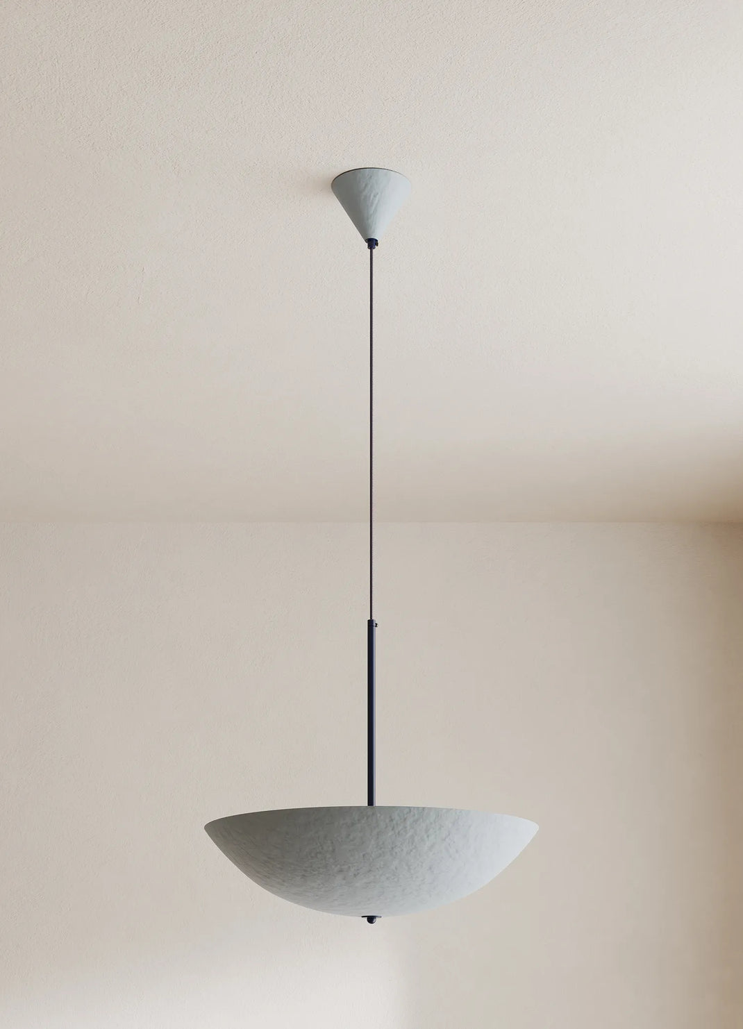 A modern pendant light with a textured, bowl-shaped shade suspended by a thin dark blue cord from a matching ceiling canopy, set against a minimalist beige wall and ceiling.
