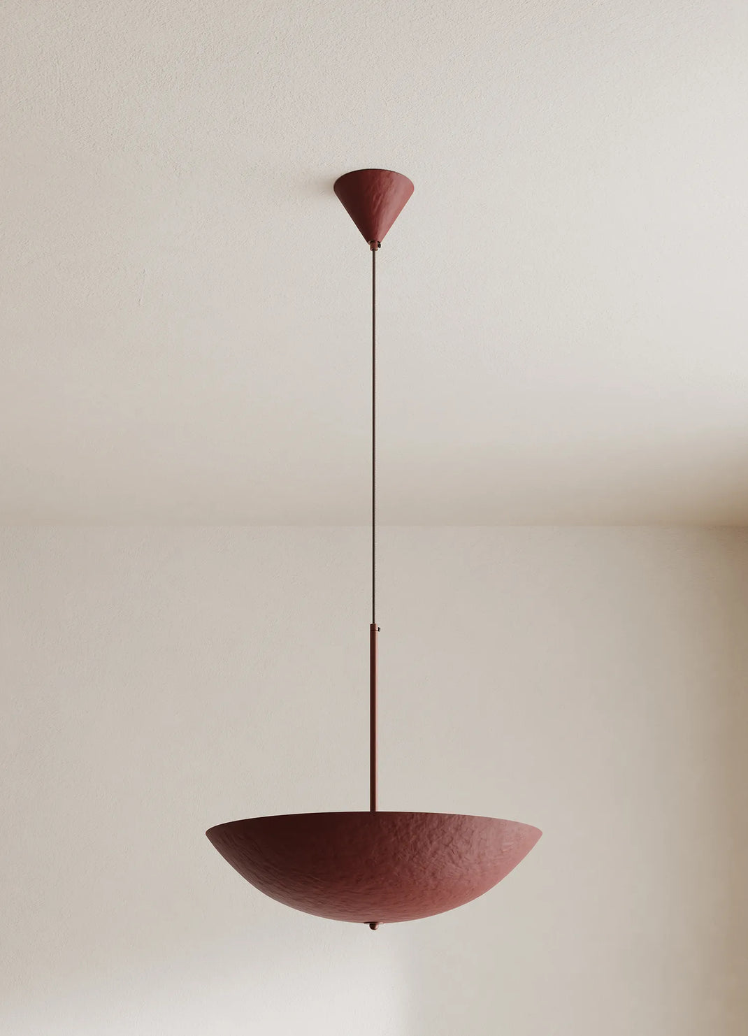 A modern pendant light with a textured deep red-colored bowl shade and matching ceiling canopy, suspended by a thin rod against a neutral off-white wall.