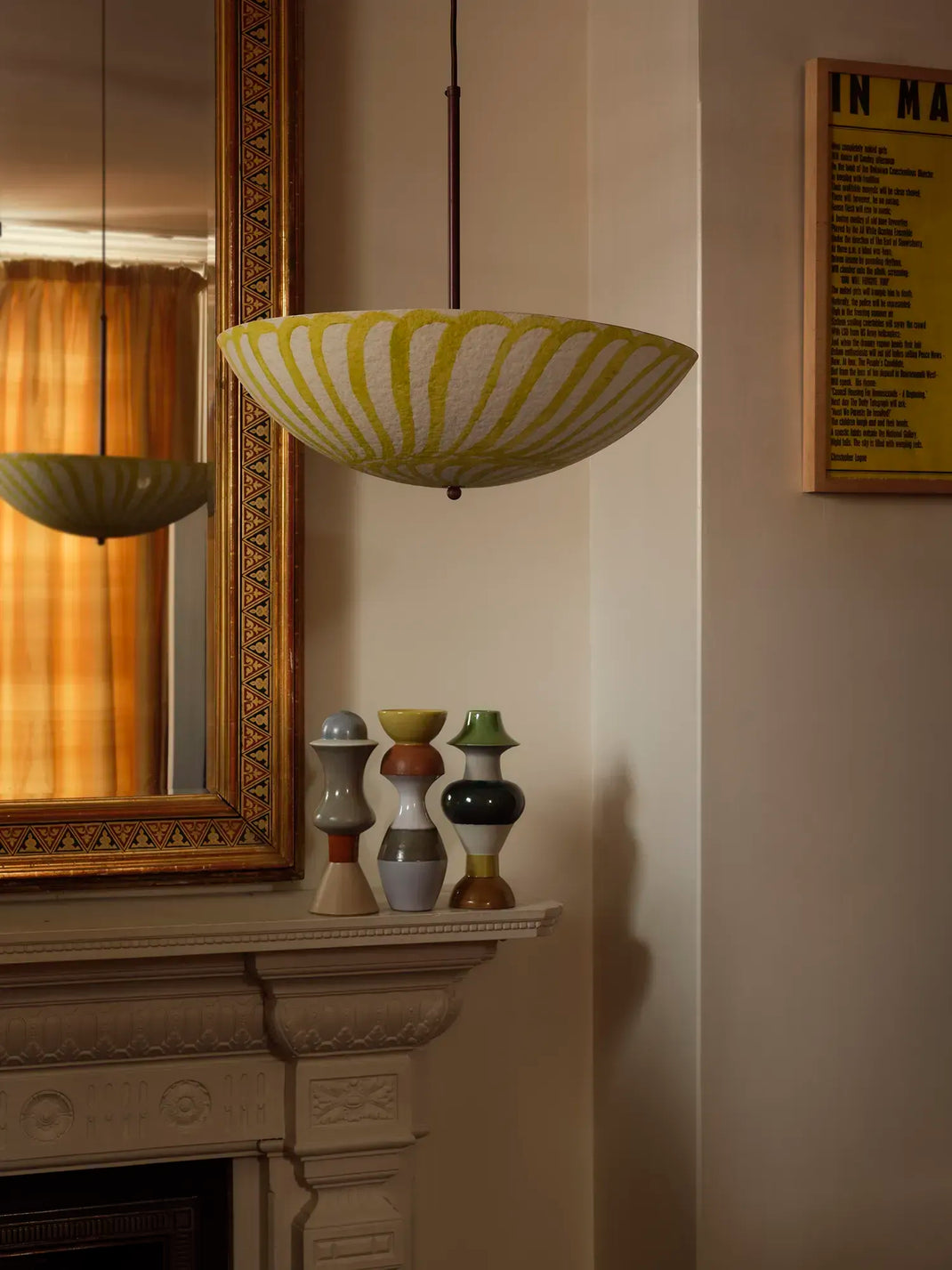A yellow and white striped bowl-shaped pendant light hangs above a fireplace mantel in a warm, dimly lit room. The scene includes a gold-framed mirror reflecting the light, small decorative vases on the mantel, and a framed print on the wall.