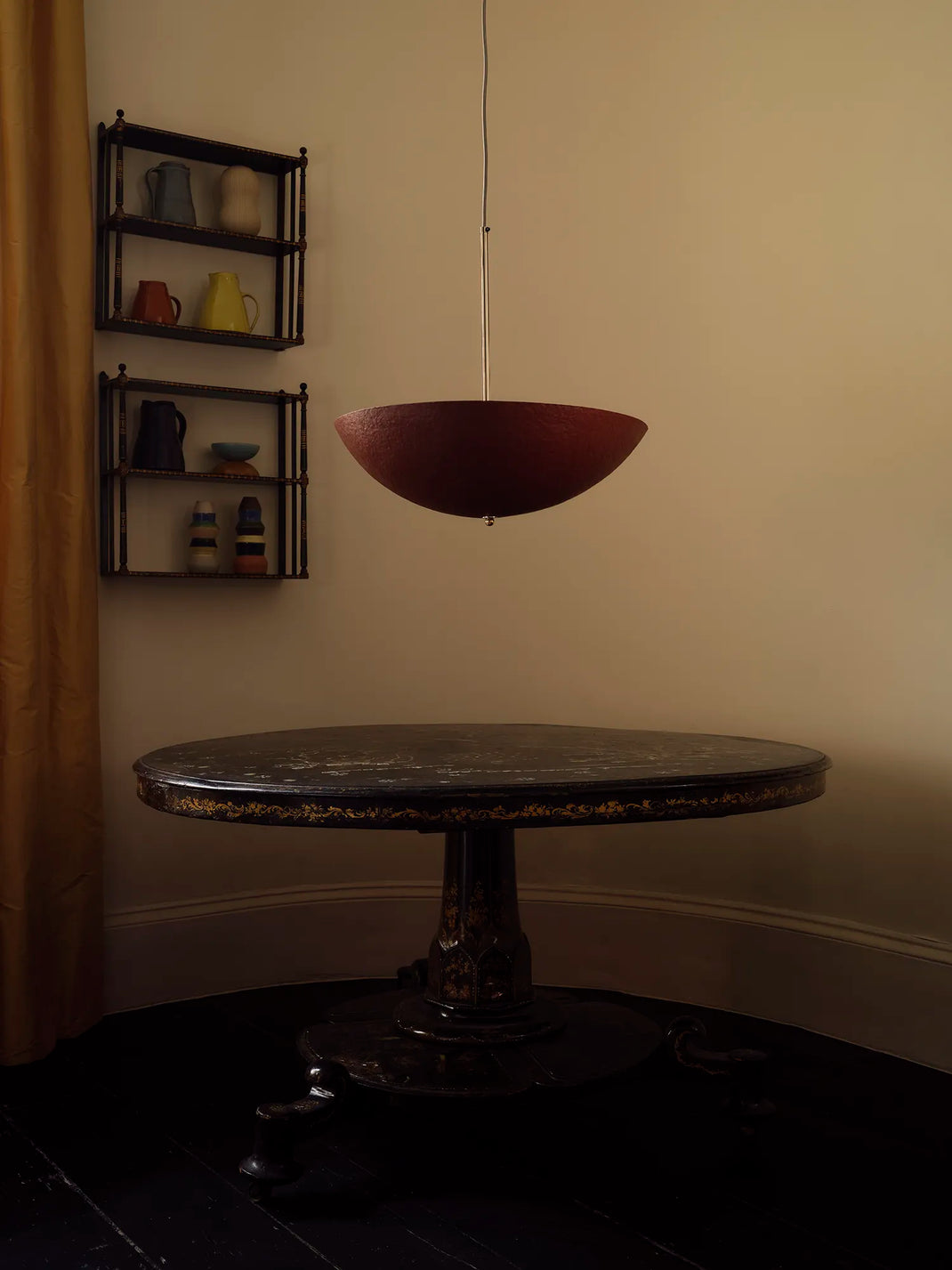 A mid-century modern pendant light with a deep red bowl-shaped shade hangs over a dark round table in a dimly lit room with beige walls and black floors.