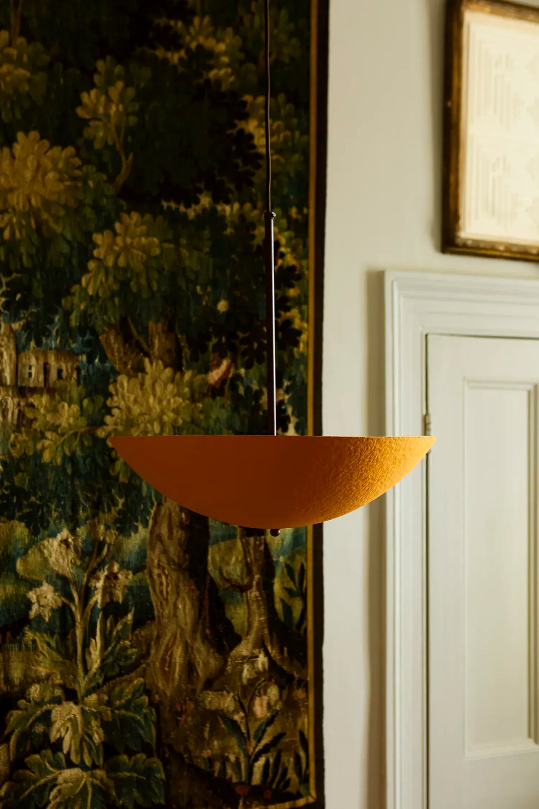 A modern, bowl-shaped pendant light in a textured terracotta orange finish, suspended by a thin black cord in an elegant interior setting with a dark forest tapestry and white paneled door in the background.