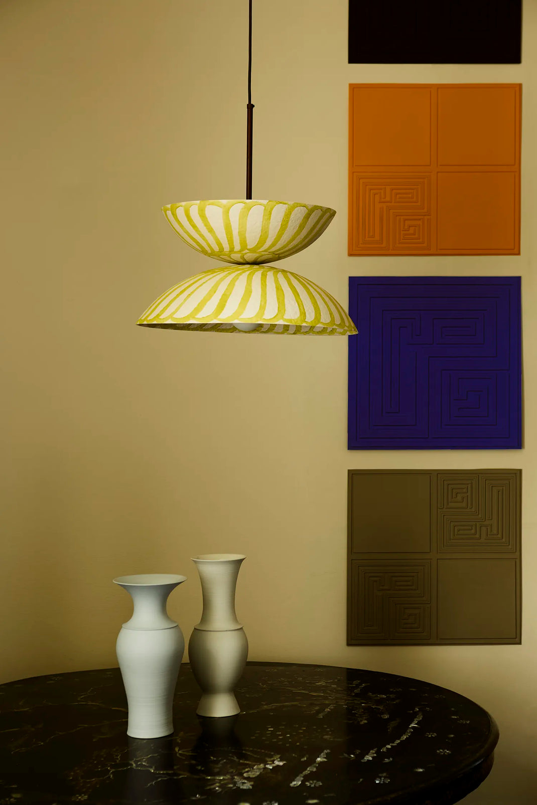 A modern two-tiered pendant light with a yellow and white pattern hangs over a dark marble table with two vases. The background wall is beige and features four colorful square panels with geometric patterns.