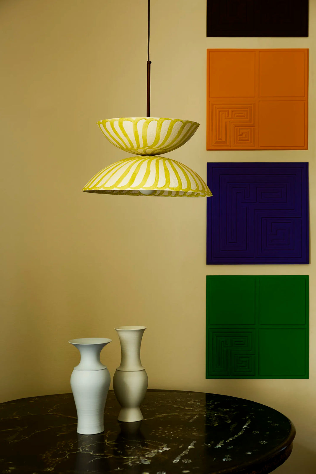 A modern two-tiered pendant light with yellow and white shades hangs over a dark marble table with two vases. The background is a beige wall decorated with four colorful square panels in orange, purple, green, and black.