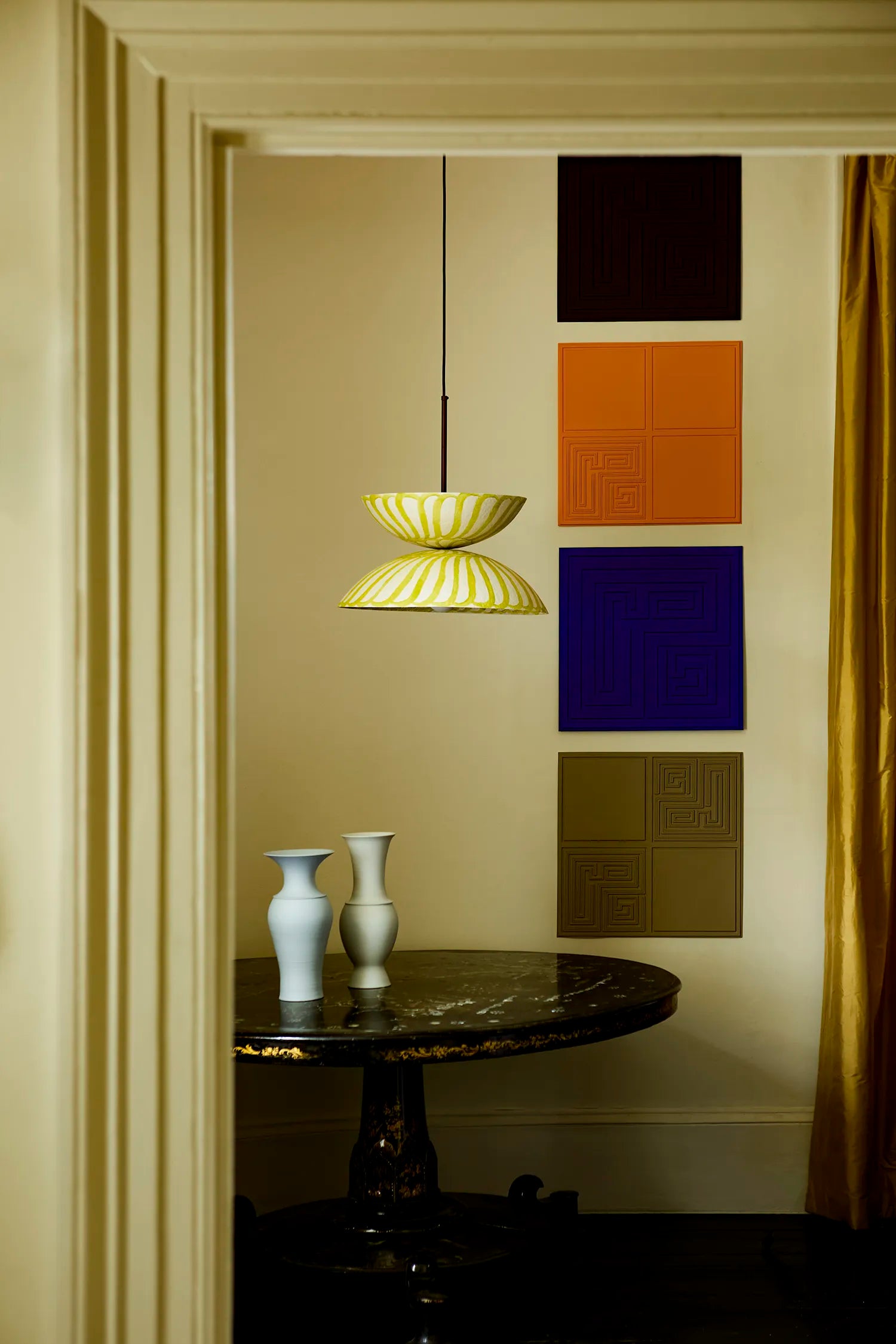 A yellow and white pendant light hangs in a room viewed through a doorway, with a dark round table, two white vases, and a wall of colorful square panels in the background.