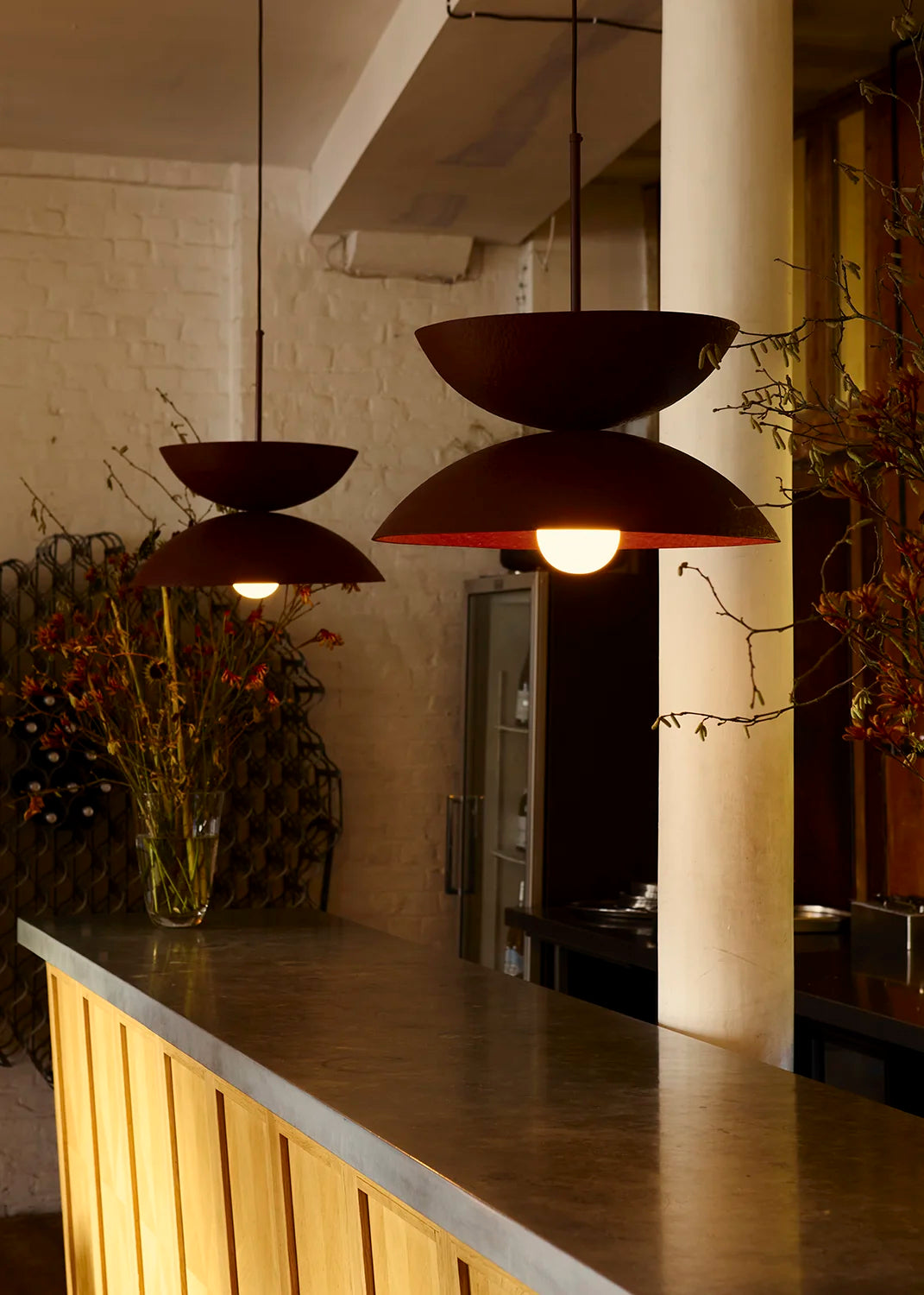 A set of two modern pendant lights with dark red-colored bowl-shaped shades hanging over a wooden bar counter in a warmly lit restaurant or cafe setting.
