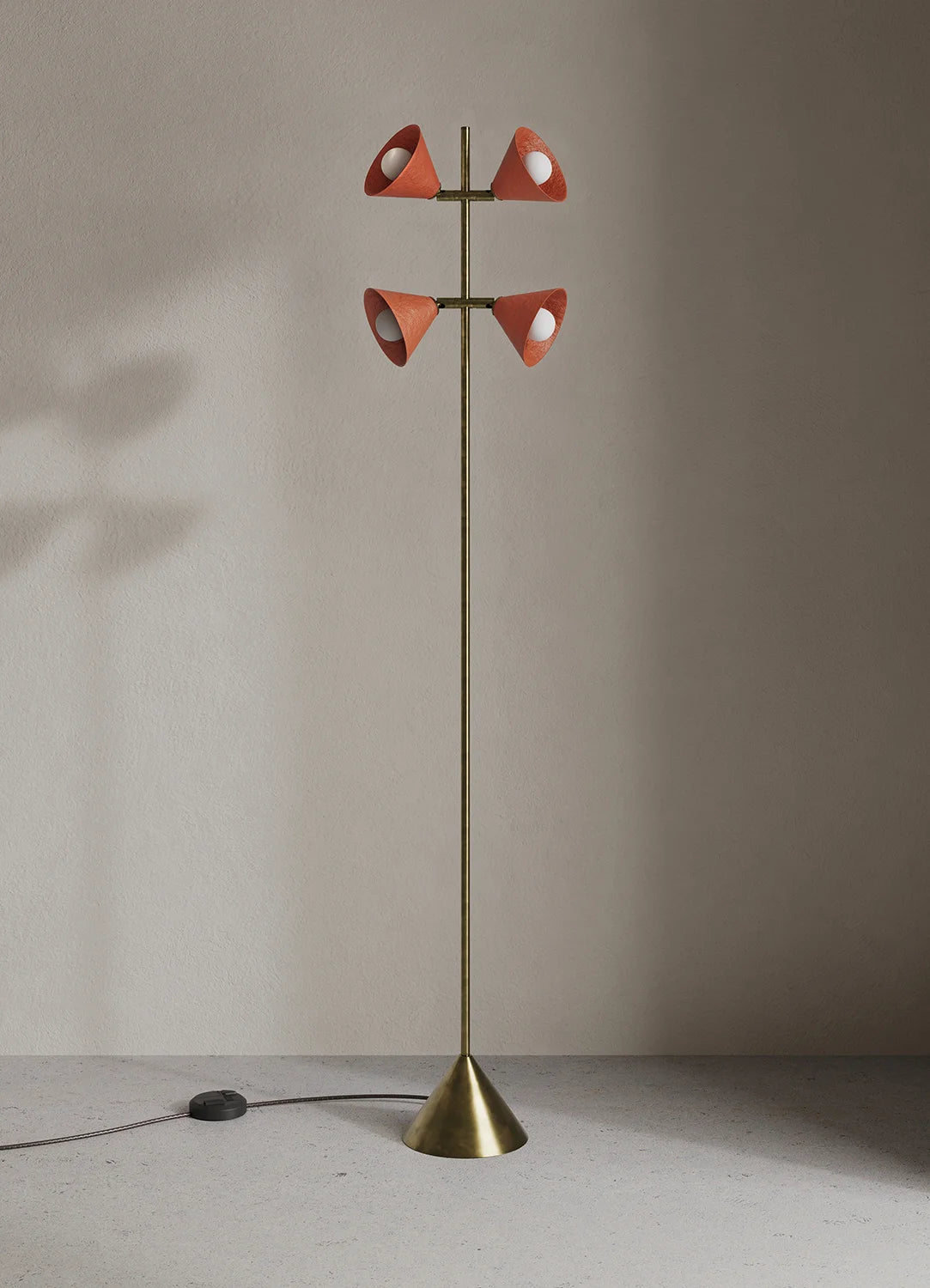 A mid-century modern floor lamp with a slim brass pole, a conical base, and four upward-facing cone shades in terracotta orange set against a neutral beige wall and floor.