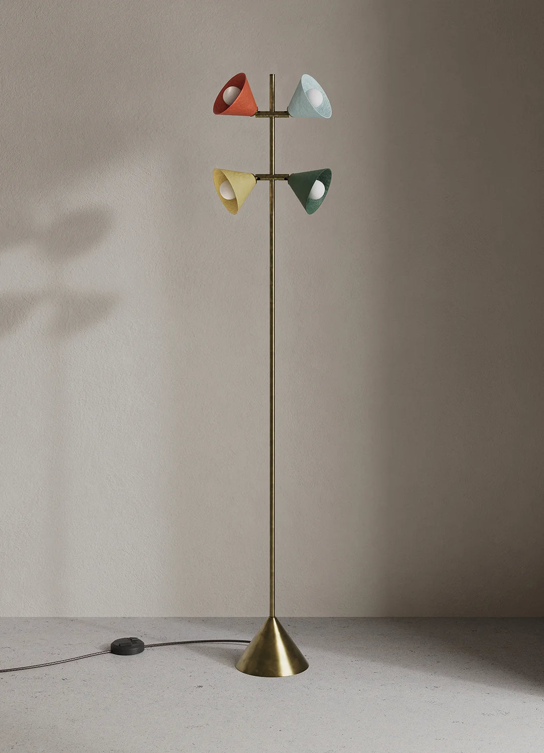 A mid-century modern floor lamp with a slim brass pole and four cone-shaped shades in red, light blue, yellow, and green, set against a neutral beige wall.