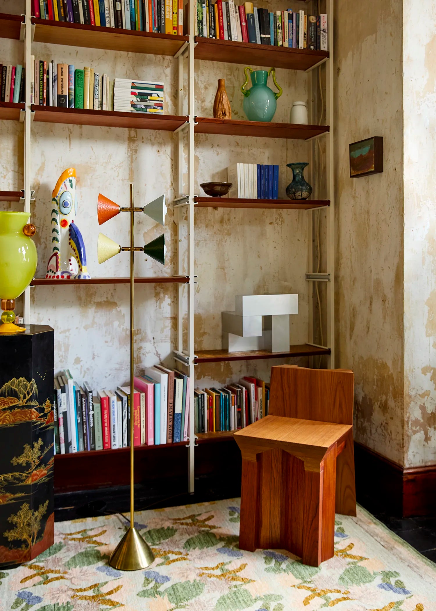 A styled interior corner featuring a wooden stool, a brass floor lamp, bookshelves filled with books, and a patterned rug against a textured wall.
