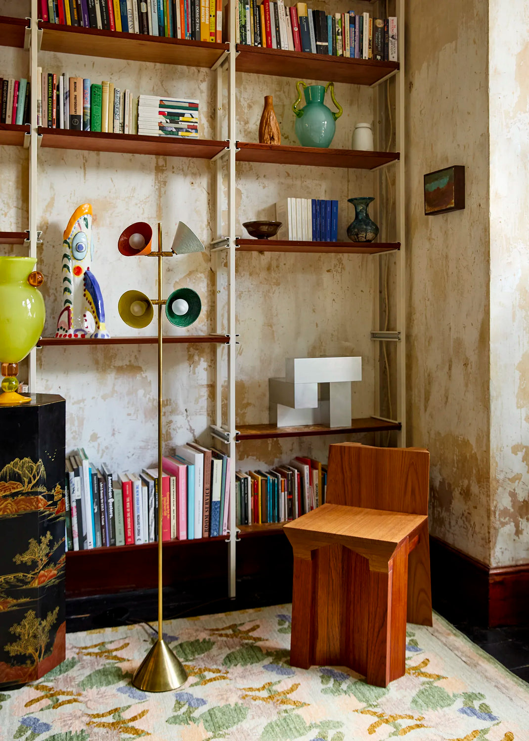 A styled interior corner featuring a wooden stool in the foreground, a tall floor lamp, open bookshelves filled with books and decorative objects, and a patterned rug against a textured wall.