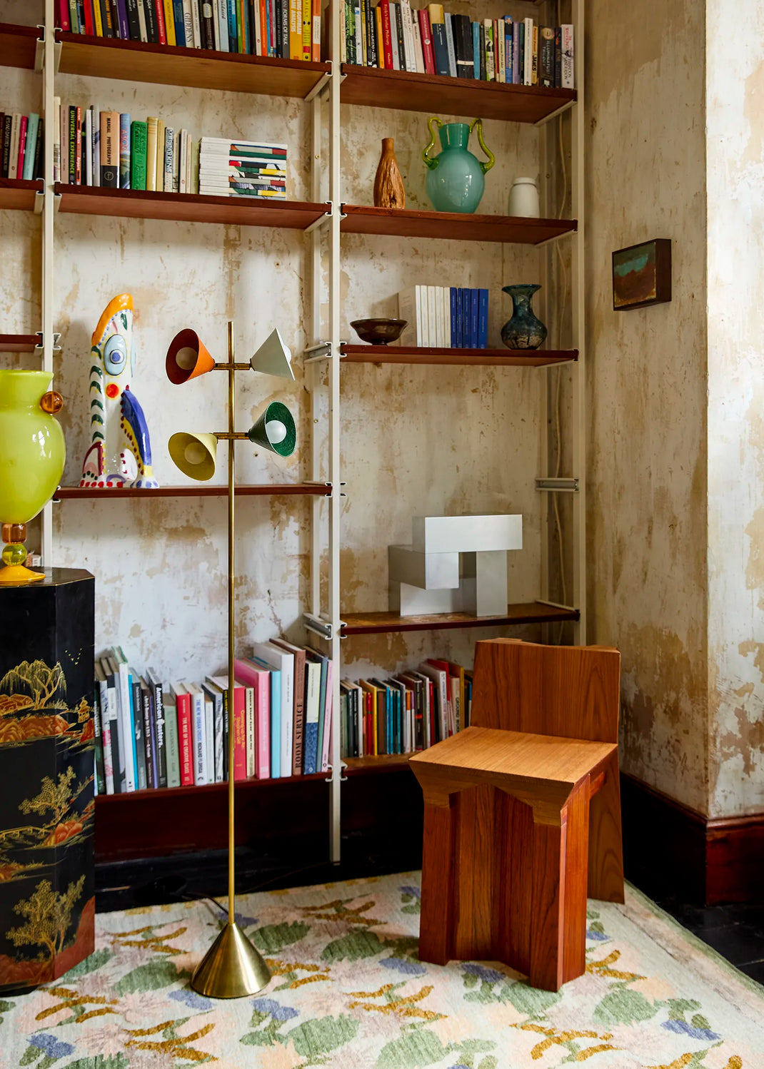 A styled interior corner featuring a brass floor lamp with multiple colorful shades, a small wooden stool, and bookshelves filled with books in a vintage room setting.