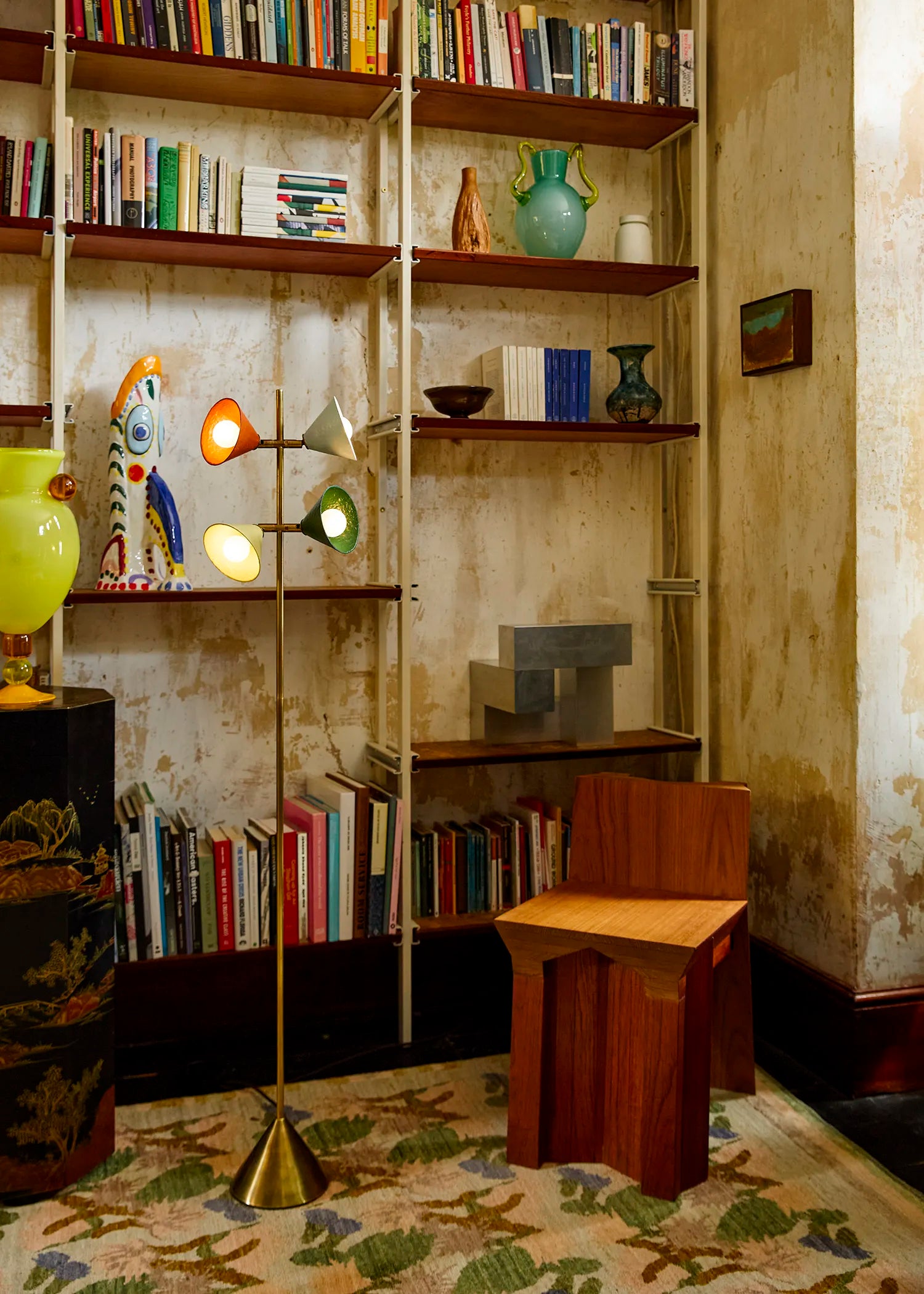 A mid-century modern floor lamp with multiple glowing orange and green cone-shaped lights stands in a vintage, book-filled room next to a wooden chair and bookshelves.