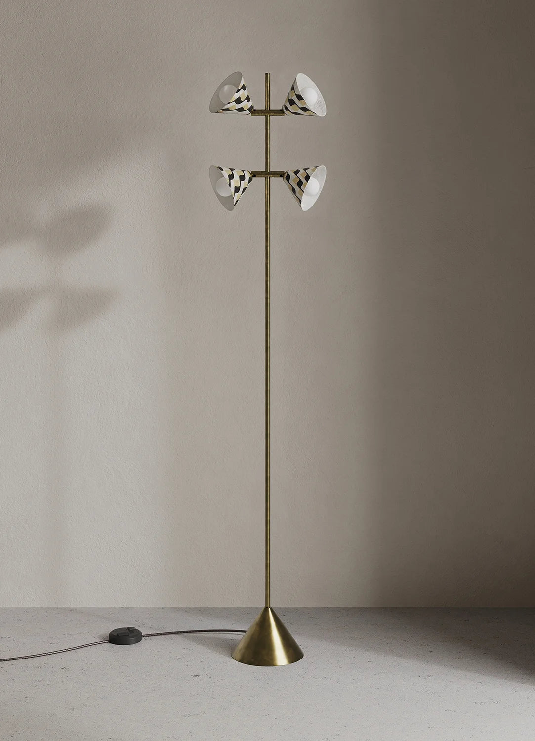 A tall brass floor lamp with four white and black patterned shades, standing on a concrete floor against a neutral beige wall.