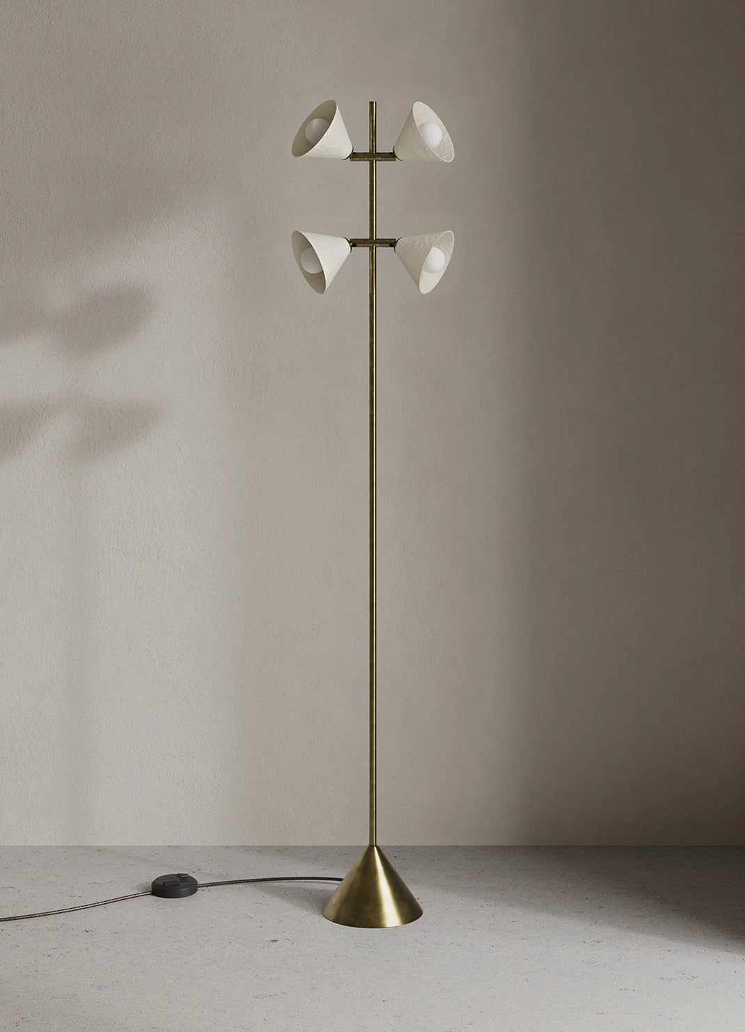 A modern floor lamp with a slim brass-colored metal stand, a conical base, and four white bell-shaped glass shades arranged in two pairs, set against a minimalist beige wall and floor.