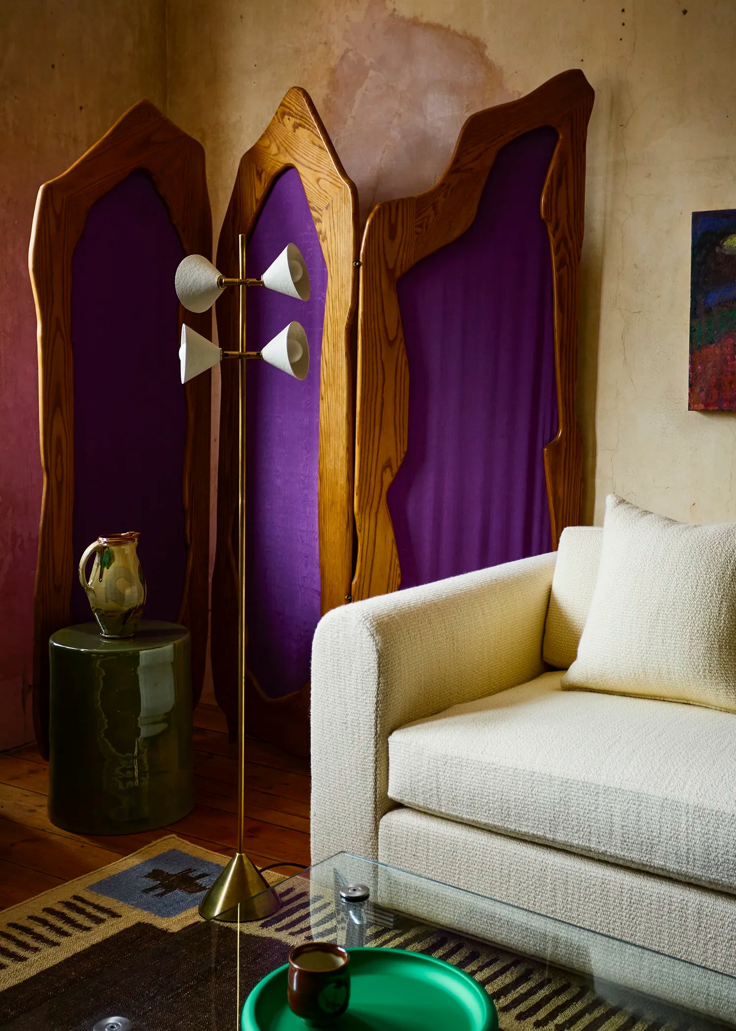 A mid-century modern brass floor lamp with white cone-shaped shades stands in a styled room next to a cream-colored sofa. The background features a large, decorative wooden screens with vibrant purple fabric panels, and a small green side table holding a ceramic pitcher.