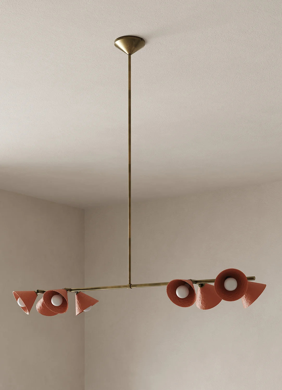 A modern linear chandelier with a brass-finished metal frame and terracotta-colored cone-shaped shades, suspended from a ceiling in a minimalist room.