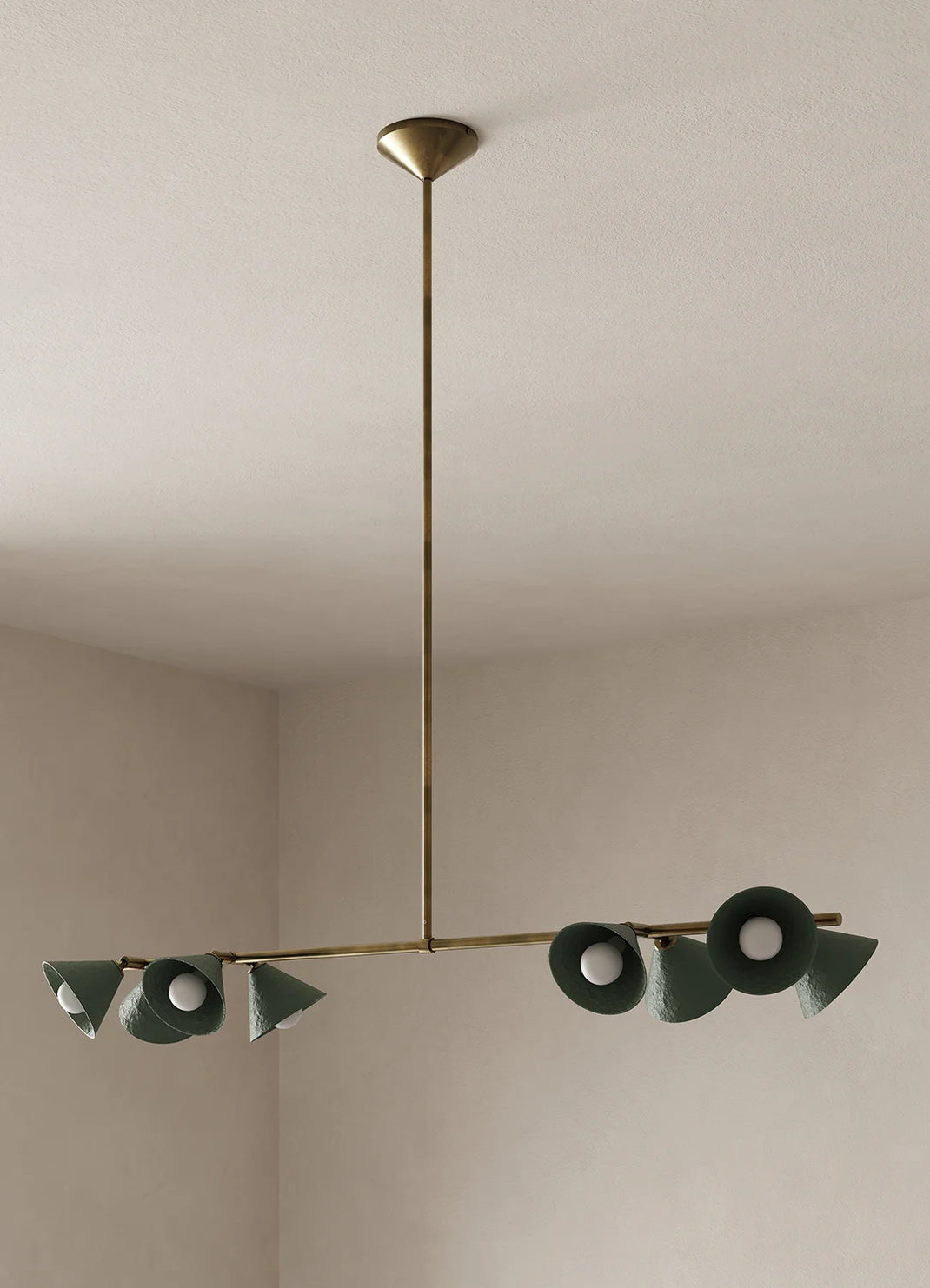 A modern brass chandelier with eight dark green ceramic cone-shaped shades, suspended from a ceiling in a minimalist room with beige walls.