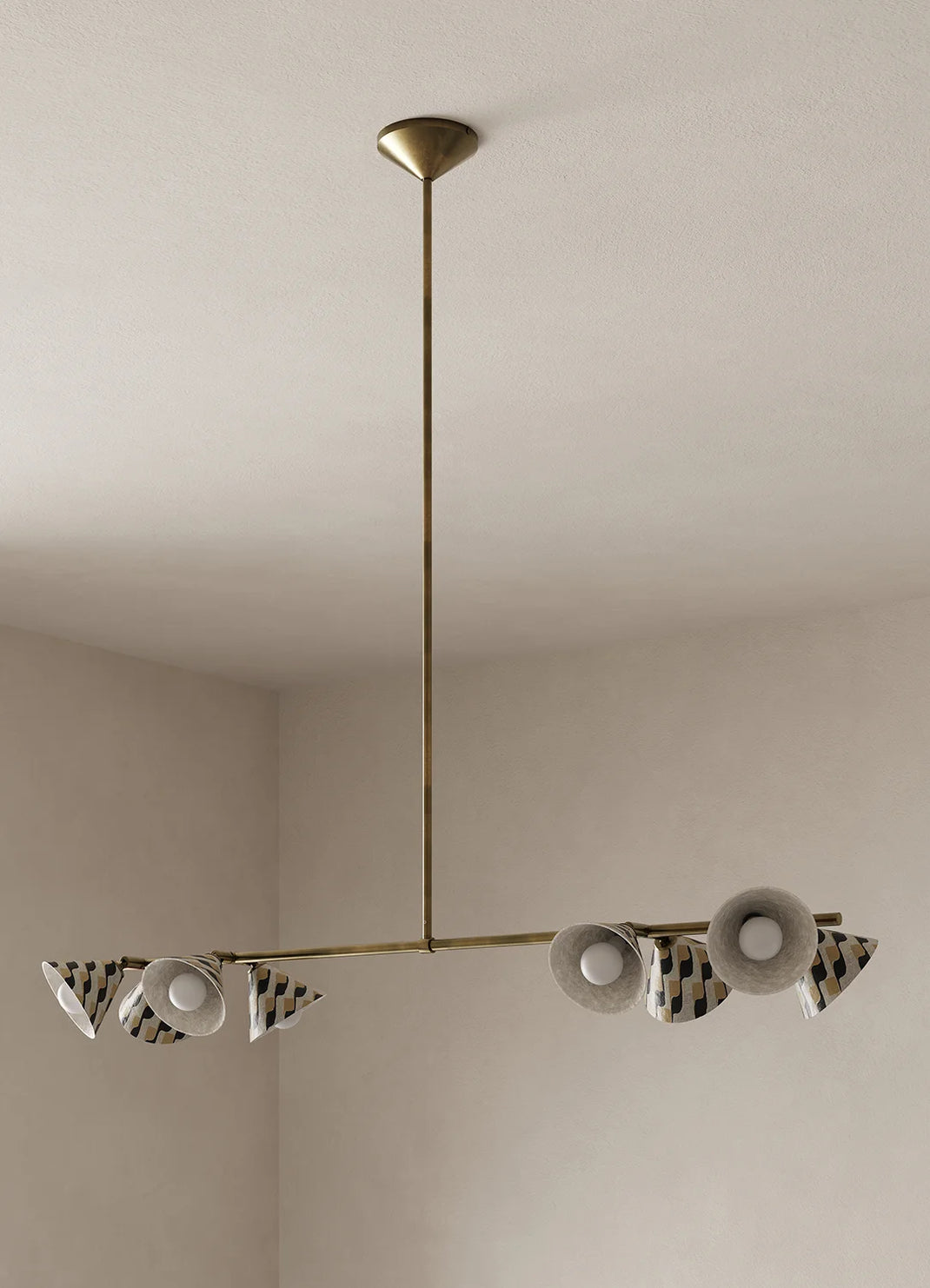 A modern brass chandelier with a central rod and two horizontal arms, featuring white and black patterned shades, shown hanging in a minimalist room with beige walls and ceiling.