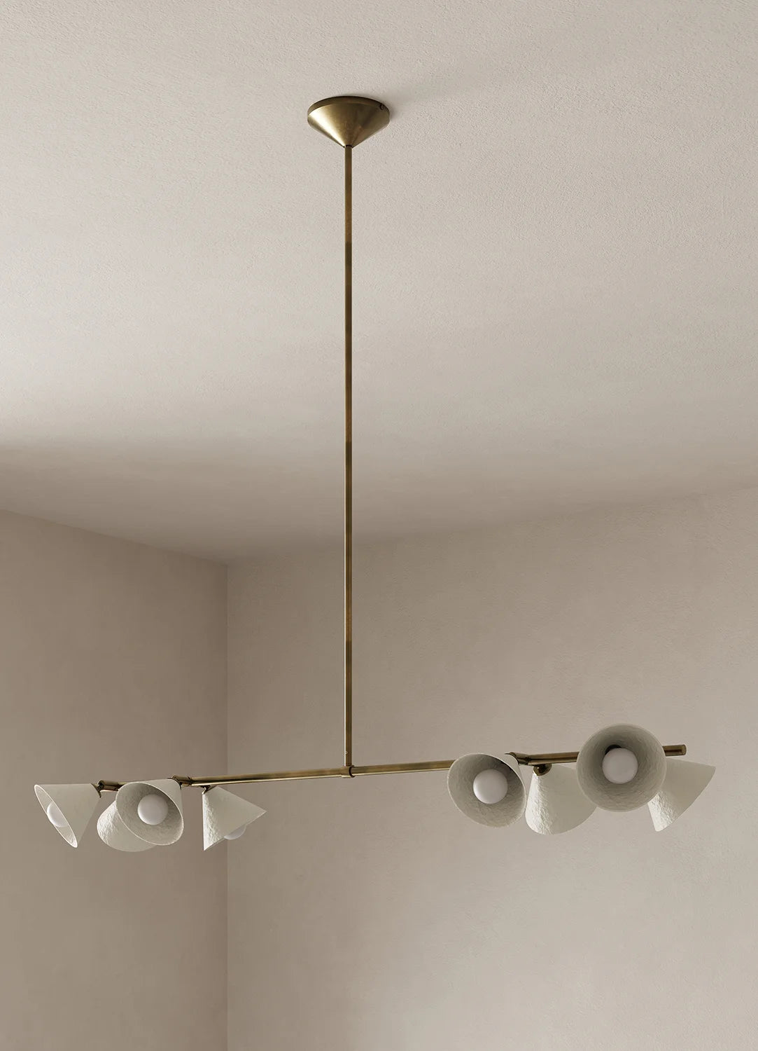 A modern linear chandelier with eight white paper pulp shades and an antique brass metal frame, suspended from a ceiling rose in a minimalist room with off-white walls.