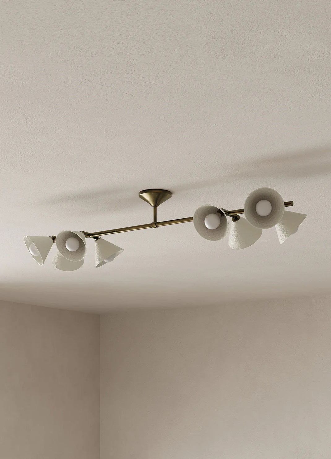 A modern linear ceiling light fixture with a brass/gold-toned central bar and eight off-white shades, mounted on a textured neutral ceiling in a minimalist room corner.