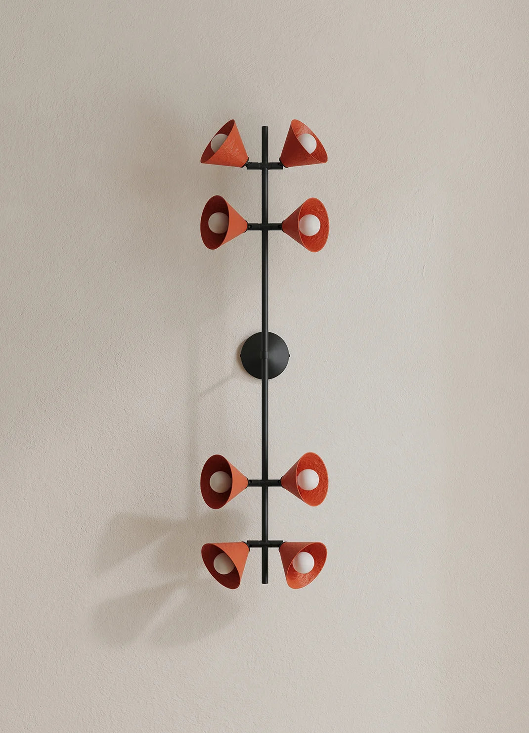 A modern wall-mounted light fixture with a black vertical frame and eight terracotta-colored cone-shaped shades arranged symmetrically, shown against a plain beige wall.