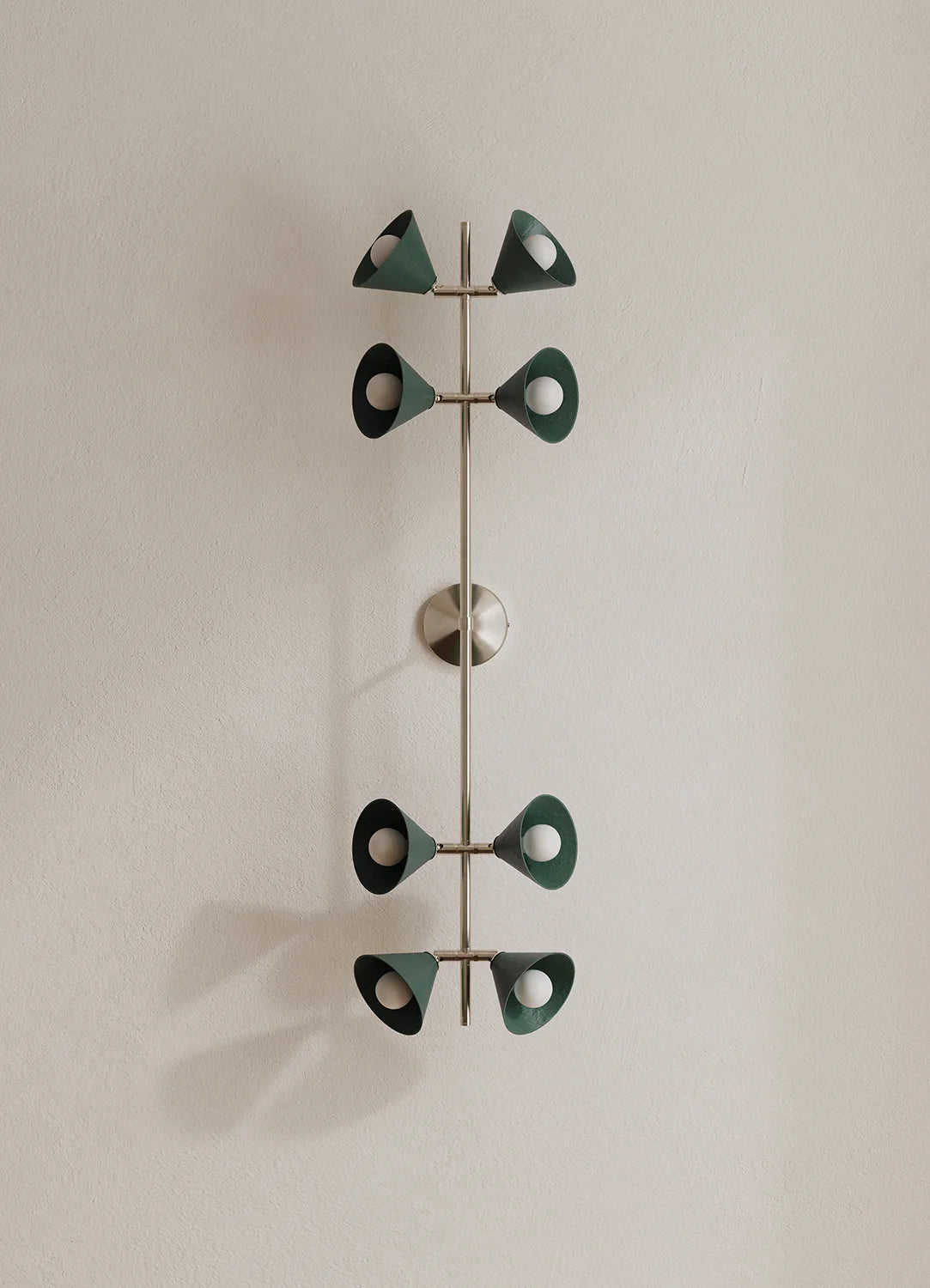 A modern wall-mounted light fixture with a vertical polished metal rod and eight decorative leaf-like shades in dark green, mounted on a plain off-white wall.