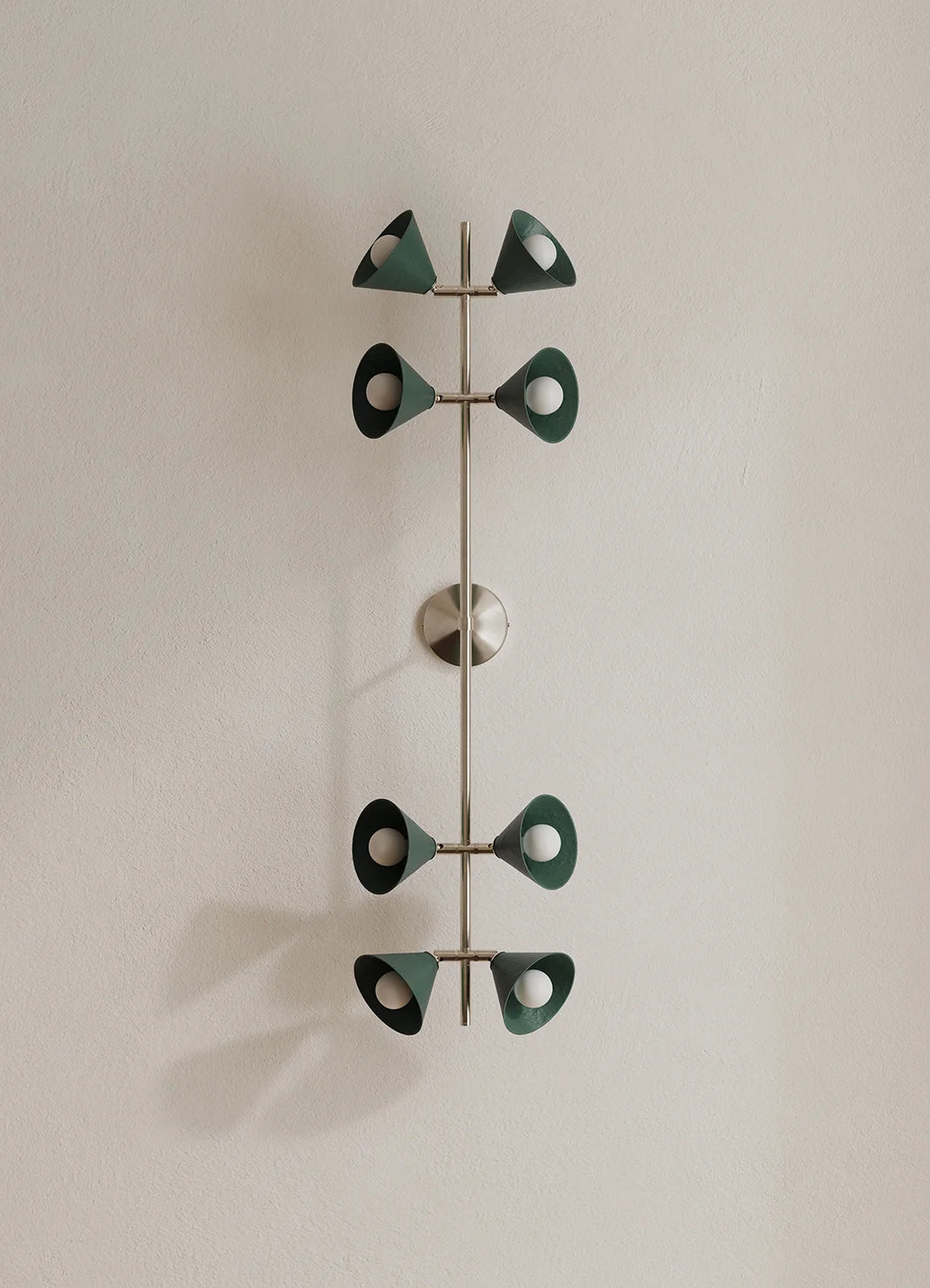 A modern wall-mounted light fixture with a vertical polished metal rod and eight decorative leaf-like shades in dark green, mounted on a plain off-white wall.