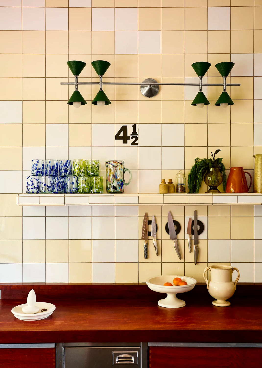 A styled kitchen interior with cream and white square tiles. A wall light with eight green cones on a polished nickel rod is mounted horizontally on the wall.