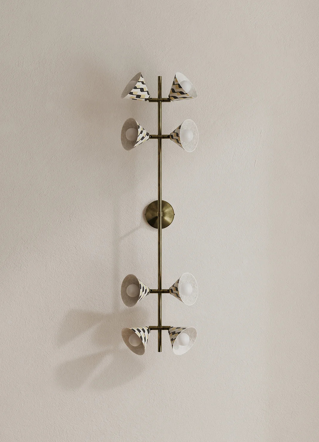 A modern wall-mounted light fixture with a vertical bronze-toned central rod and eight angled arms, each holding a white shade with a black geometric pattern, set against a plain beige wall.