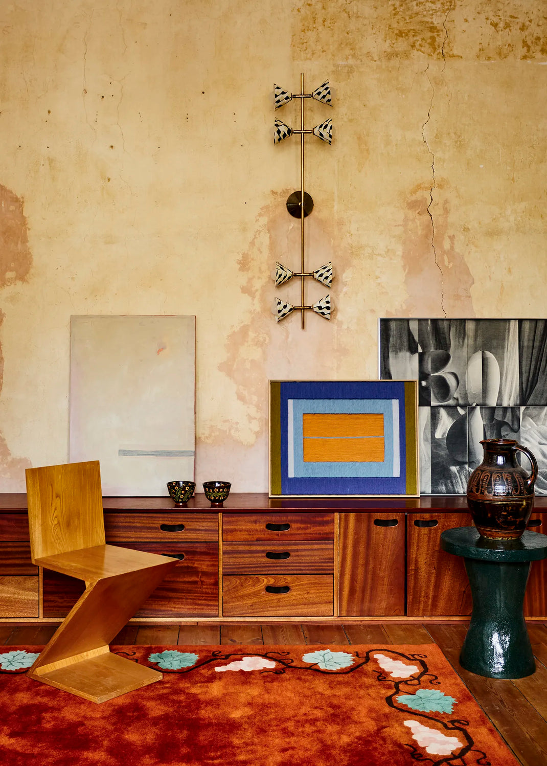 A styled interior scene with a wooden sculptural chair and a red patterned rug. A wall light with 8 shades in a white and black pattern is mounted on the wall.