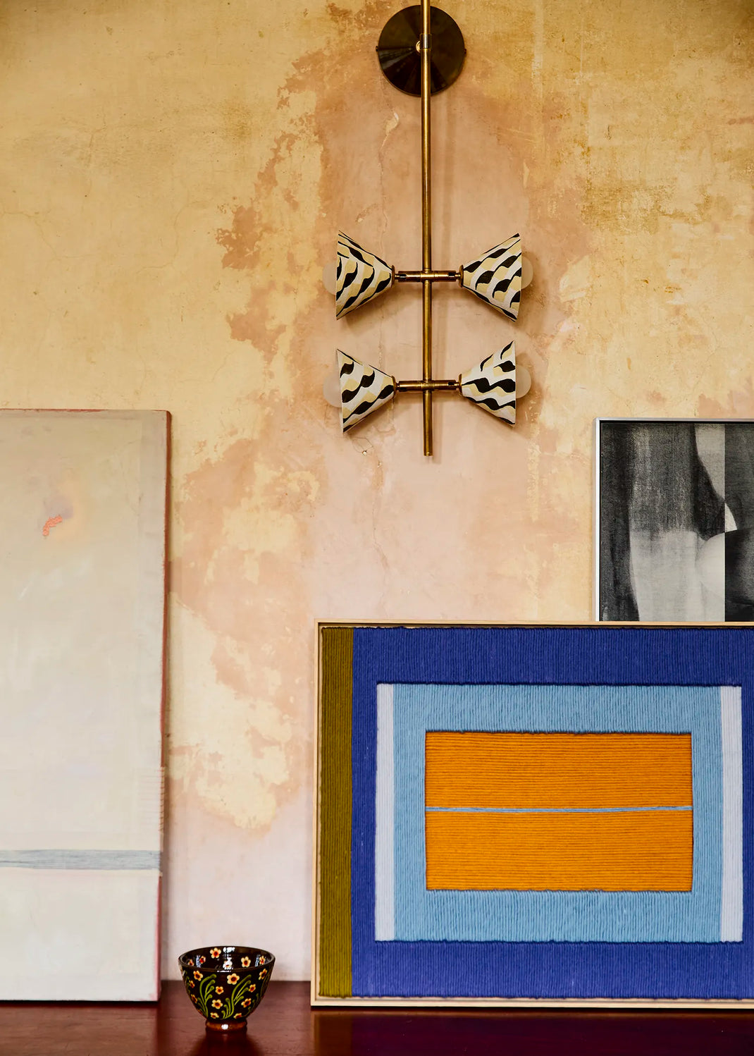 A modern wall-mounted light fixture with four bow-shaped shades in a black-and-white pattern, suspended from a brass rod on a textured beige wall. Below, framed abstract art pieces and a small decorative bowl are arranged on a dark surface.