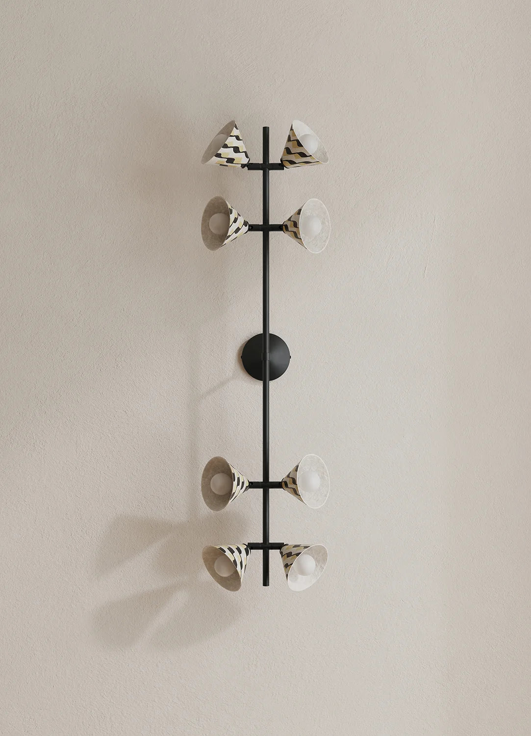 A modern wall-mounted light fixture with a vertical black metal frame and eight cone-shaped shades, some with black-and-white patterned exteriors and white interiors, mounted on a beige wall.