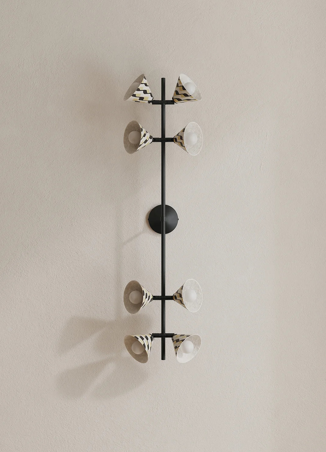 A modern wall-mounted light fixture with a vertical black metal frame and eight cone-shaped shades, some with black-and-white patterned exteriors and white interiors, mounted on a beige wall.