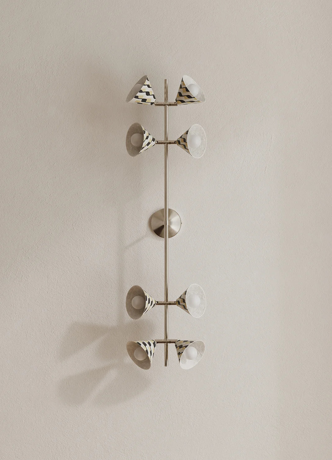 A modern wall-mounted light fixture with a vertical polished metal rod and eight angled lamp shades, some patterned with black and white stripes mounted on a beige wall.