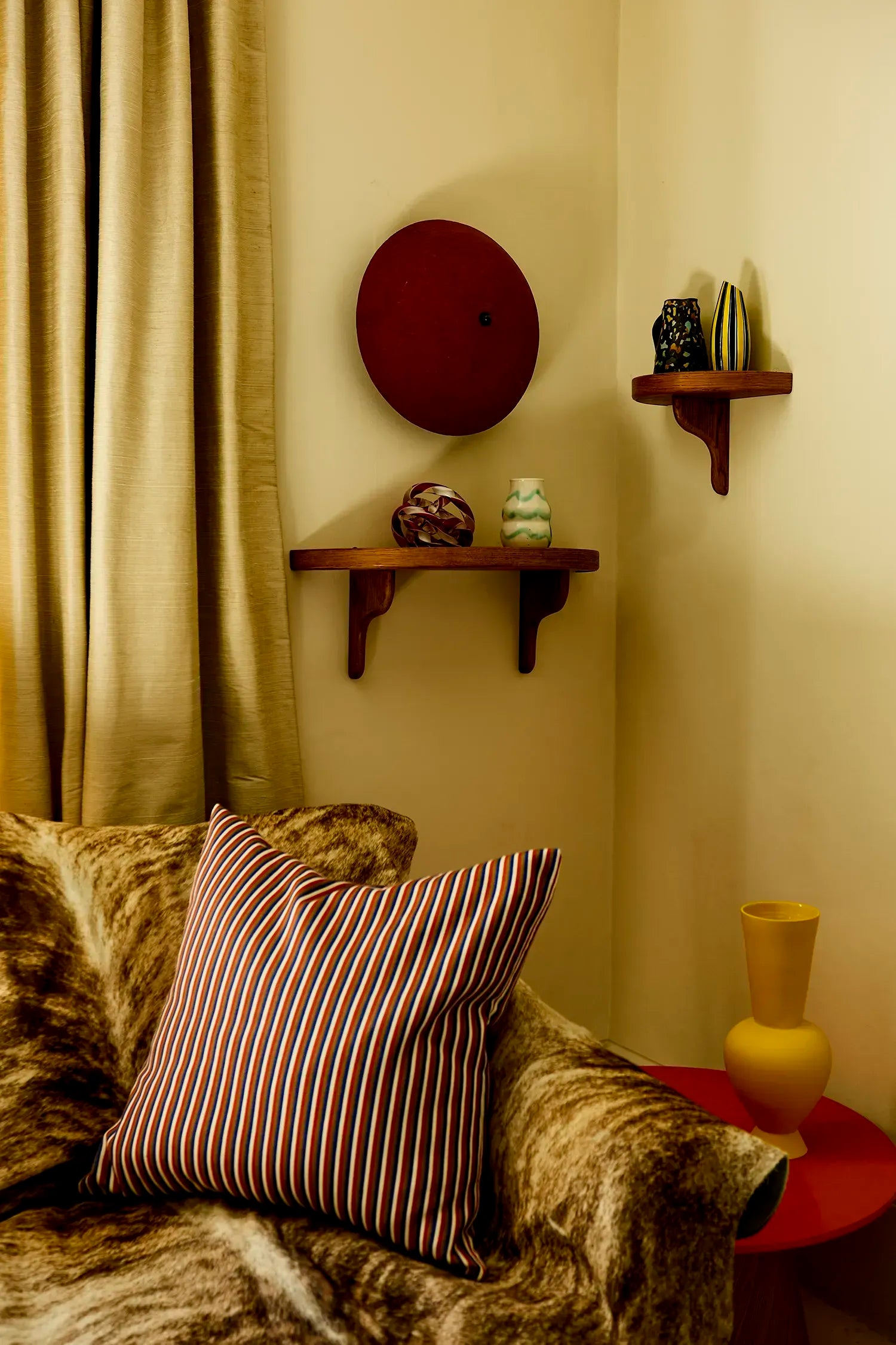 Living room corner with a striped pillow on a sofa, shelves with decorative items, and a curtain. A deep red wall sconce is mounted above the shelves.