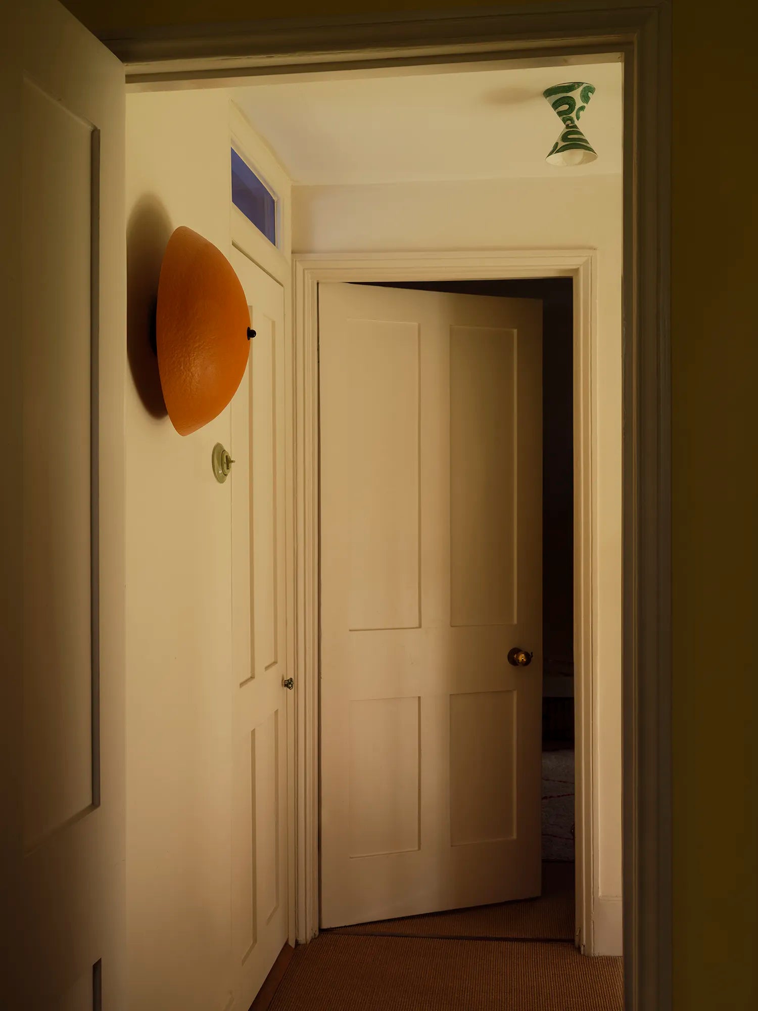 A hallway interior with cream-colored walls and doors, featuring a prominent orange wall-mounted sconce on the left and an open door in the center leading to a darker room.