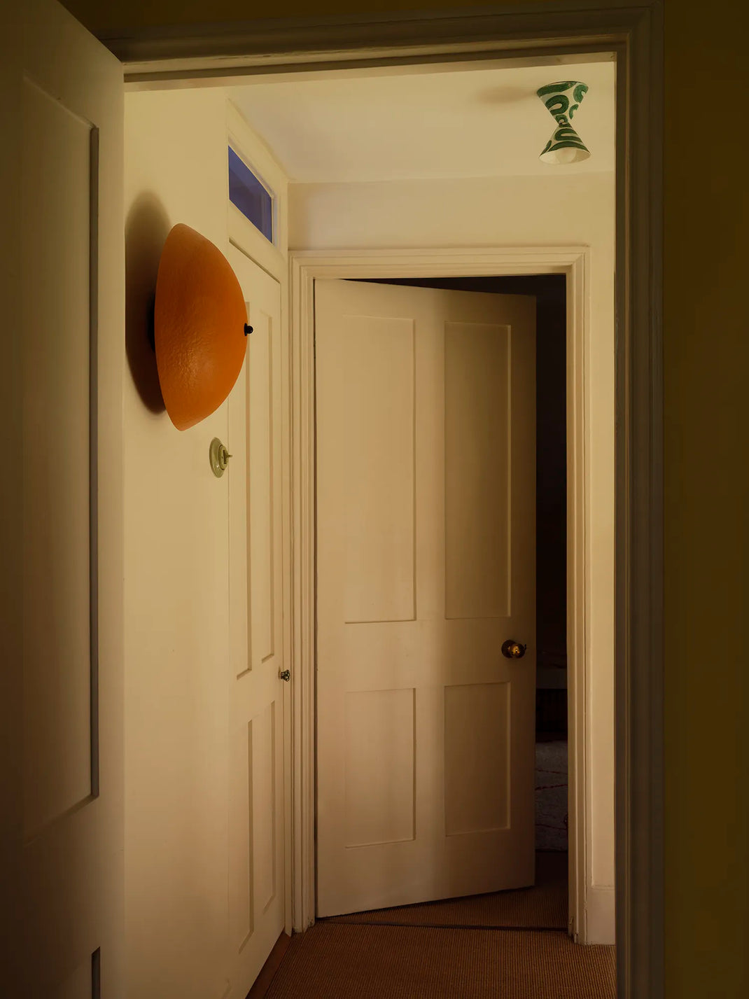 A hallway interior with cream-colored walls and doors, featuring a prominent orange wall-mounted sconce on the left and an open door in the center leading to a darker room.