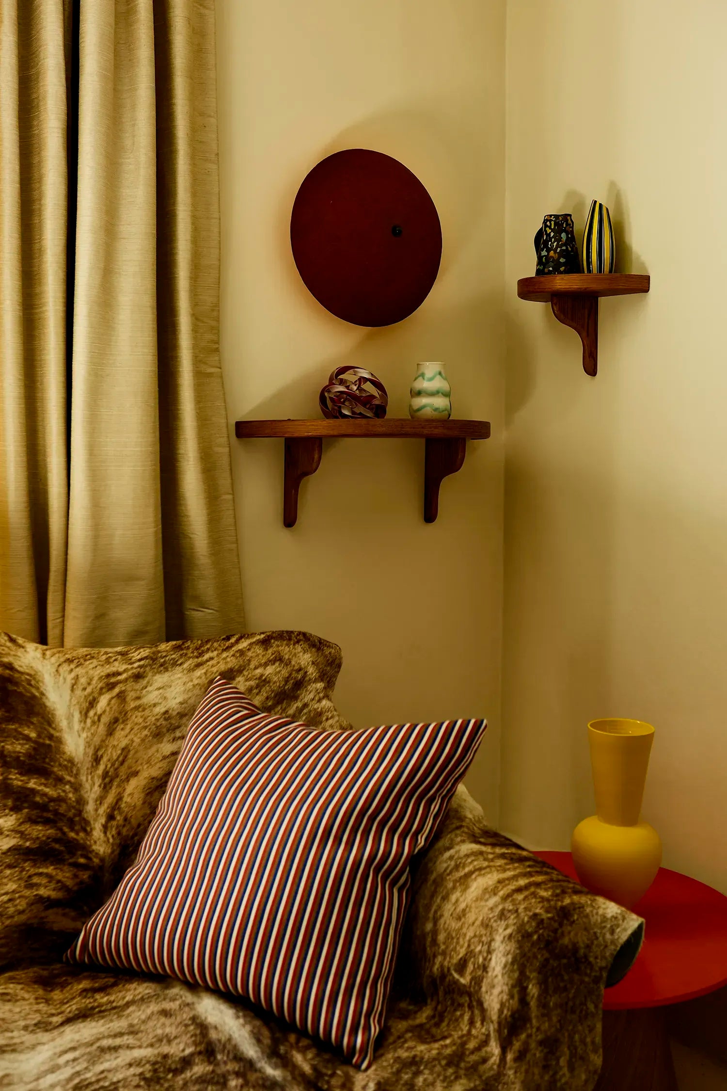 A cozy interior corner with a brown faux-fur sofa, a striped red and white throw pillow, a small red side table with a yellow vase, and wall-mounted wooden shelves holding decorative objects against a warm beige wall. A deep-red wall sconce is mounted above the wooden shelves.